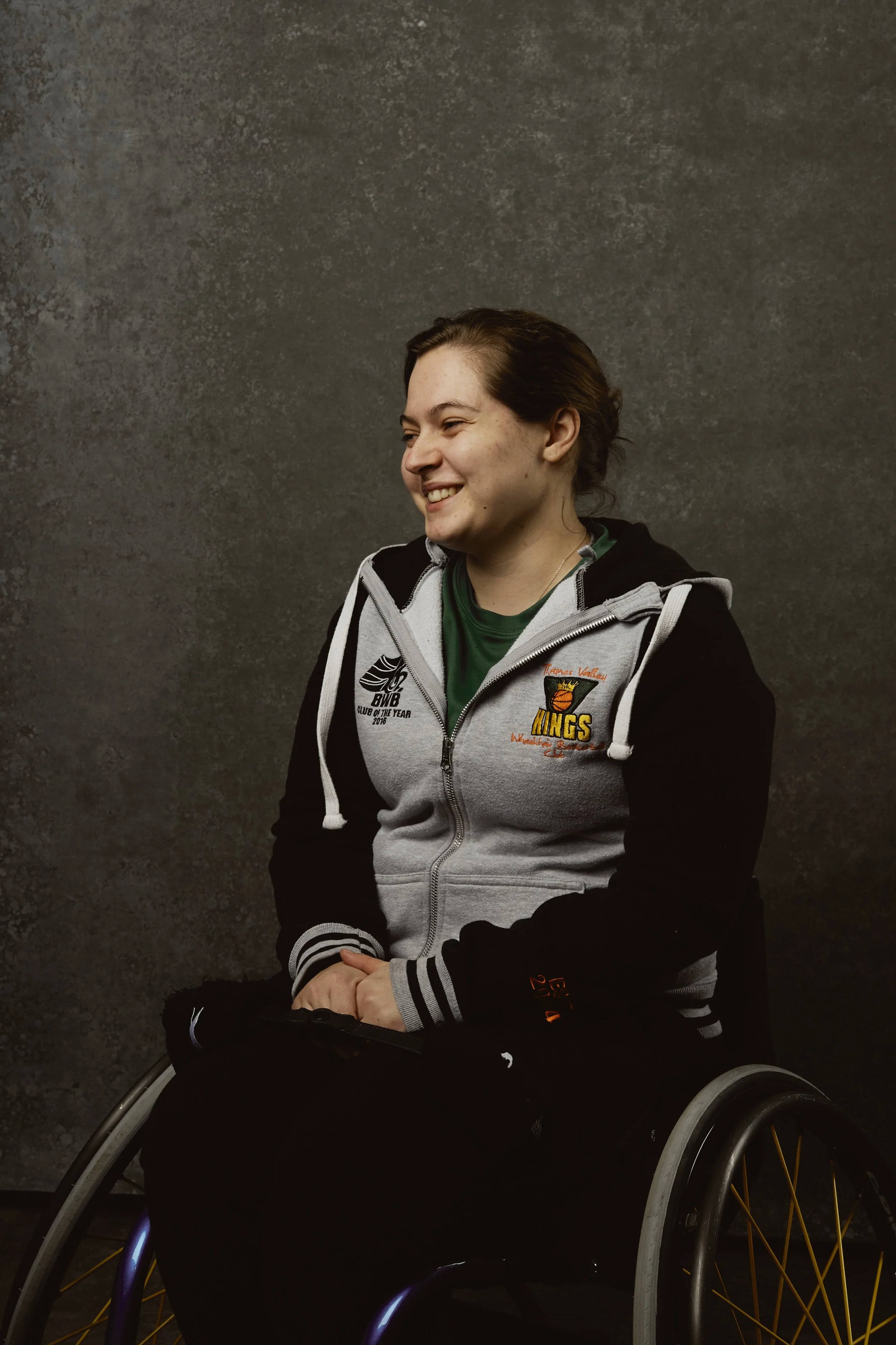 Young woman in a wheelchair smiling and facing to her right, wearing a gray and black hoodie with sports logos, against a dark textured background.