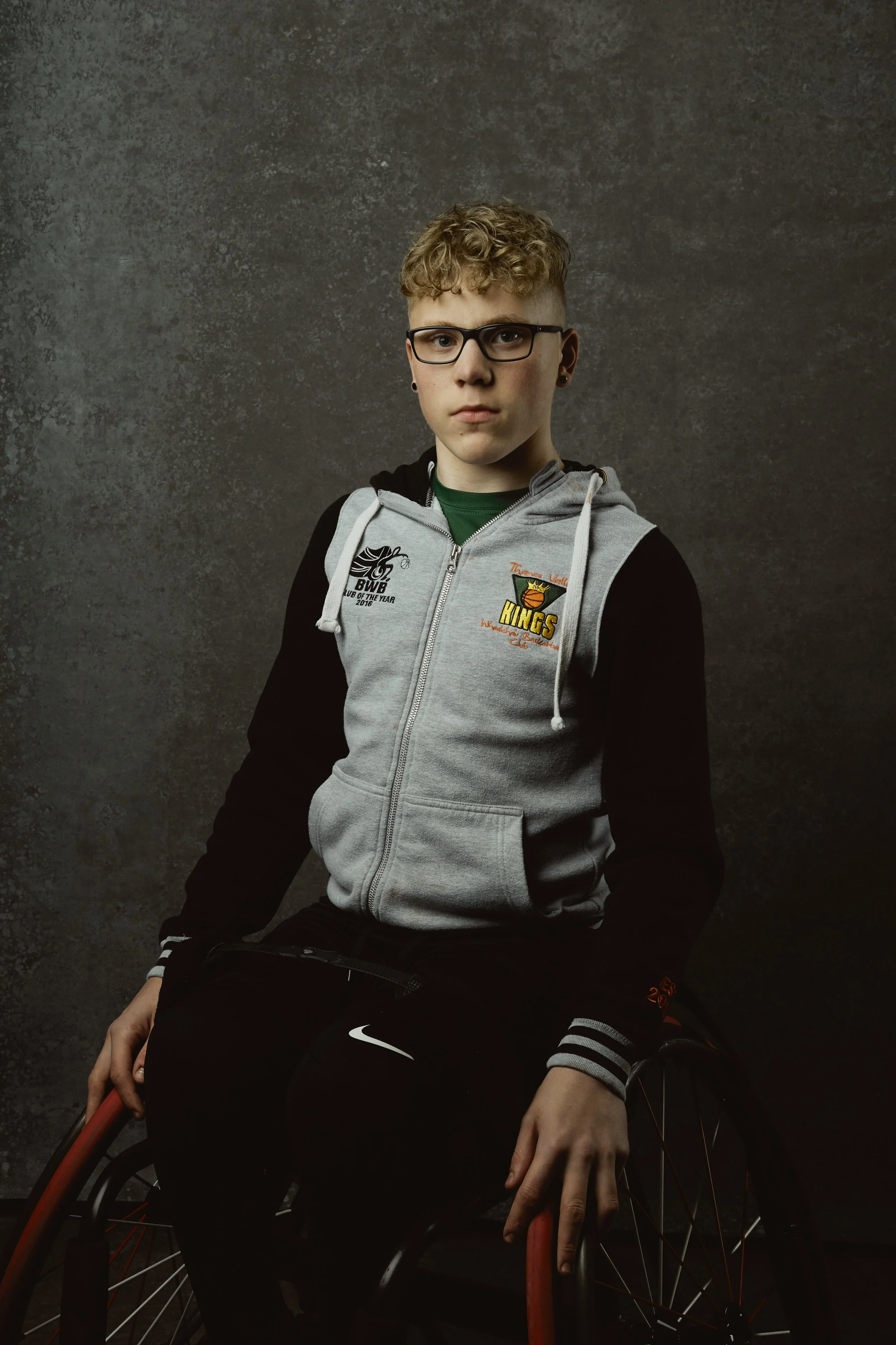 Young man with curly blonde hair, glasses, and earrings wearing a gray and black hoodie with sports logos, sitting in a wheelchair against a dark textured background.