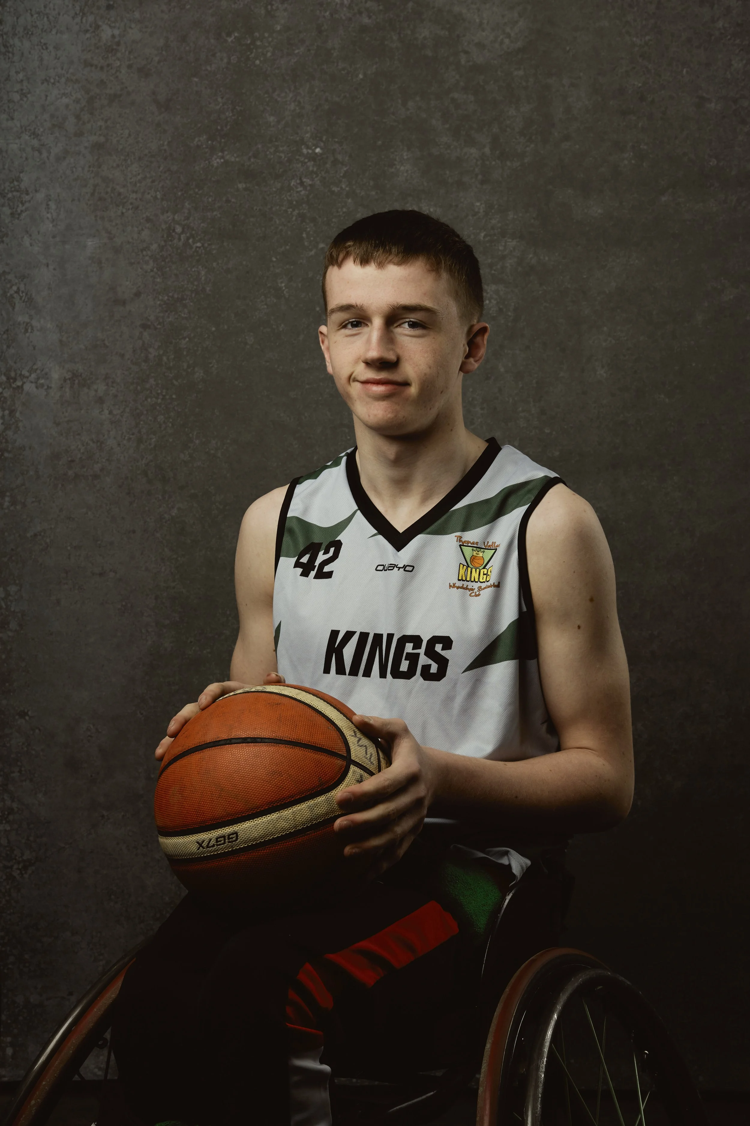 Young male wheelchair basketball player wearing a white jersey with the number 42 and 'KINGS' written on it, holding a basketball against a neutral backdrop.