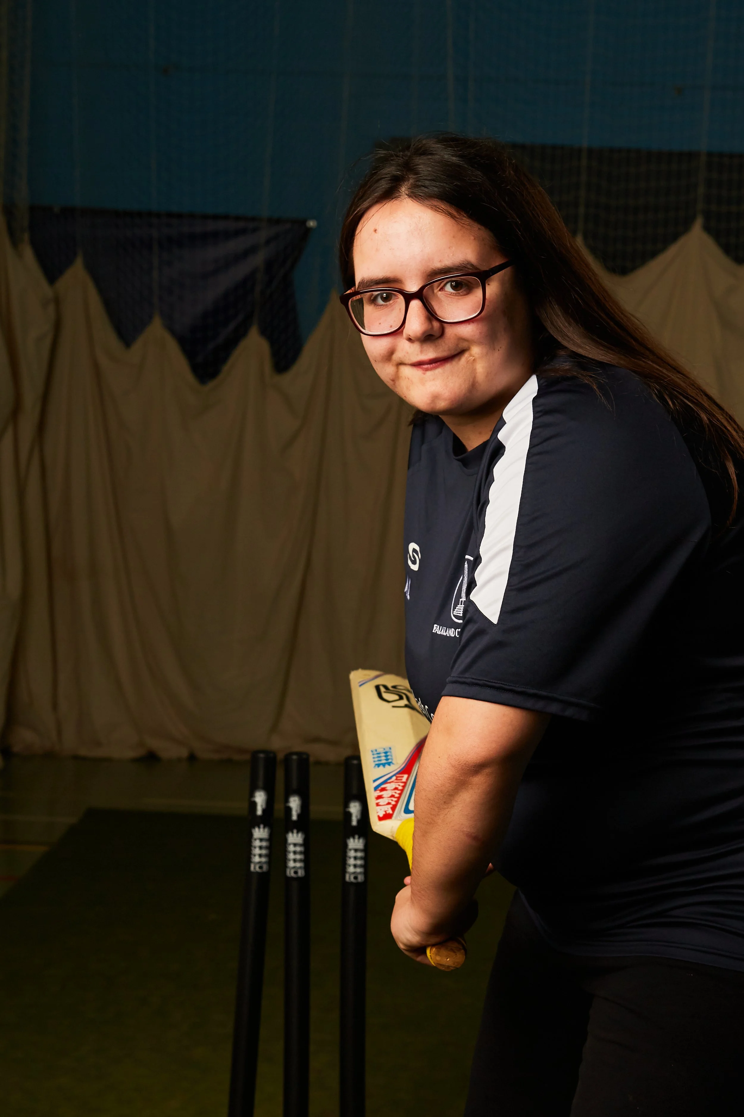 Young woman holding a cricket bat in an indoor sports venue, with cricket stumps in the background.