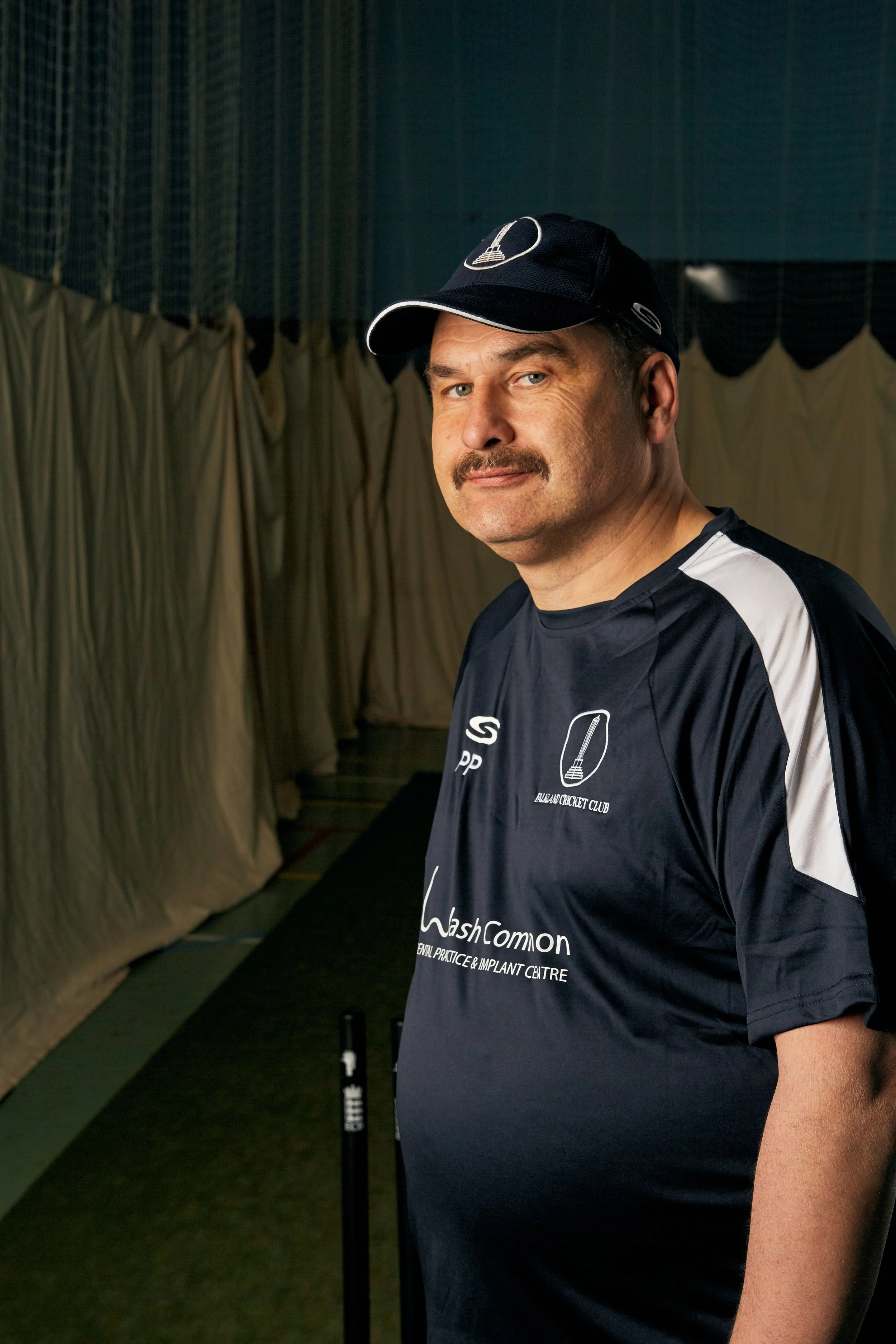 A man wearing a black sports shirt and cap posing indoors with a cricket club logo and text on his shirt.