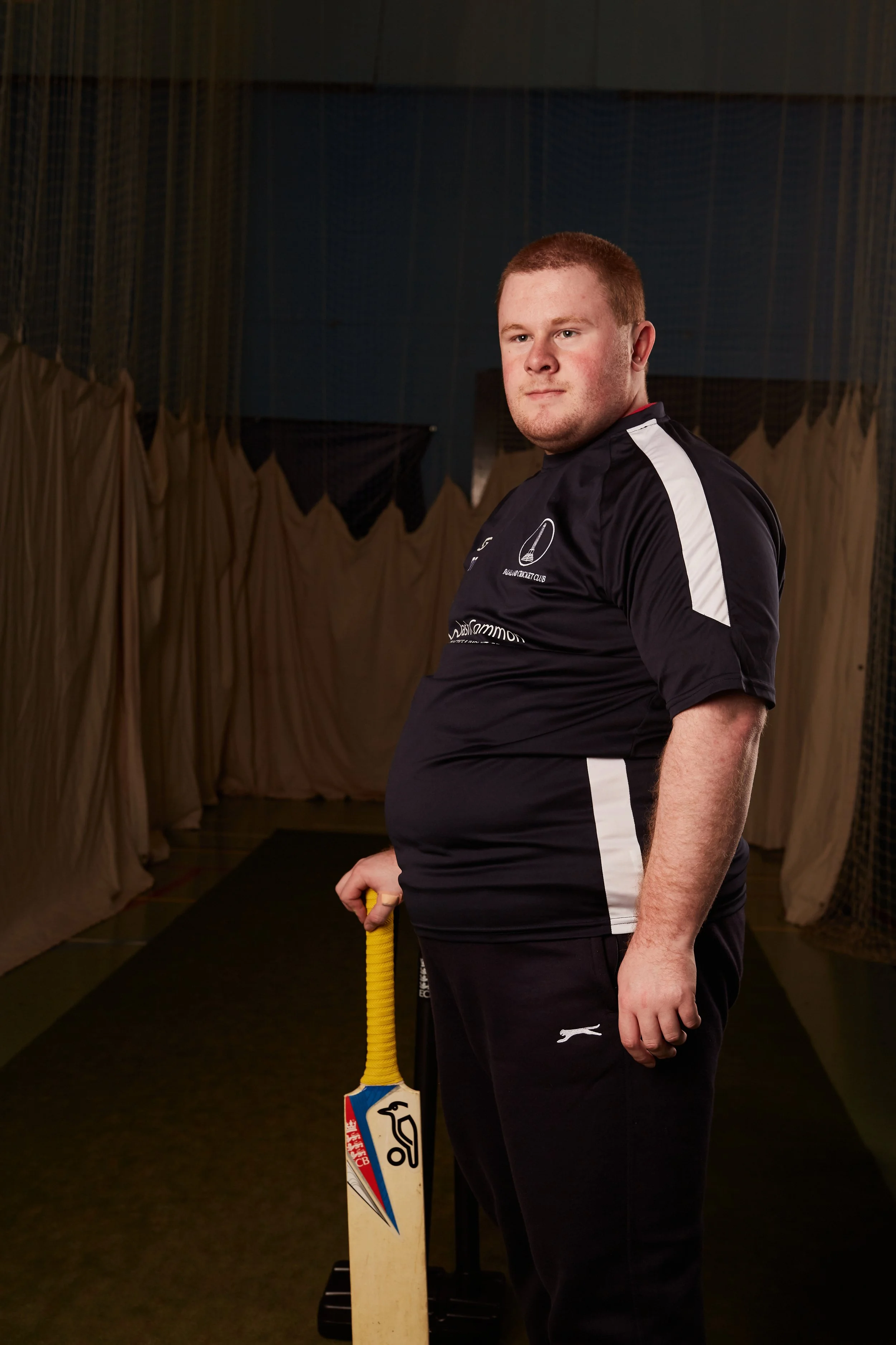A young man dressed in a black sports uniform holding a cricket bat, standing indoors with beige curtains in the background.