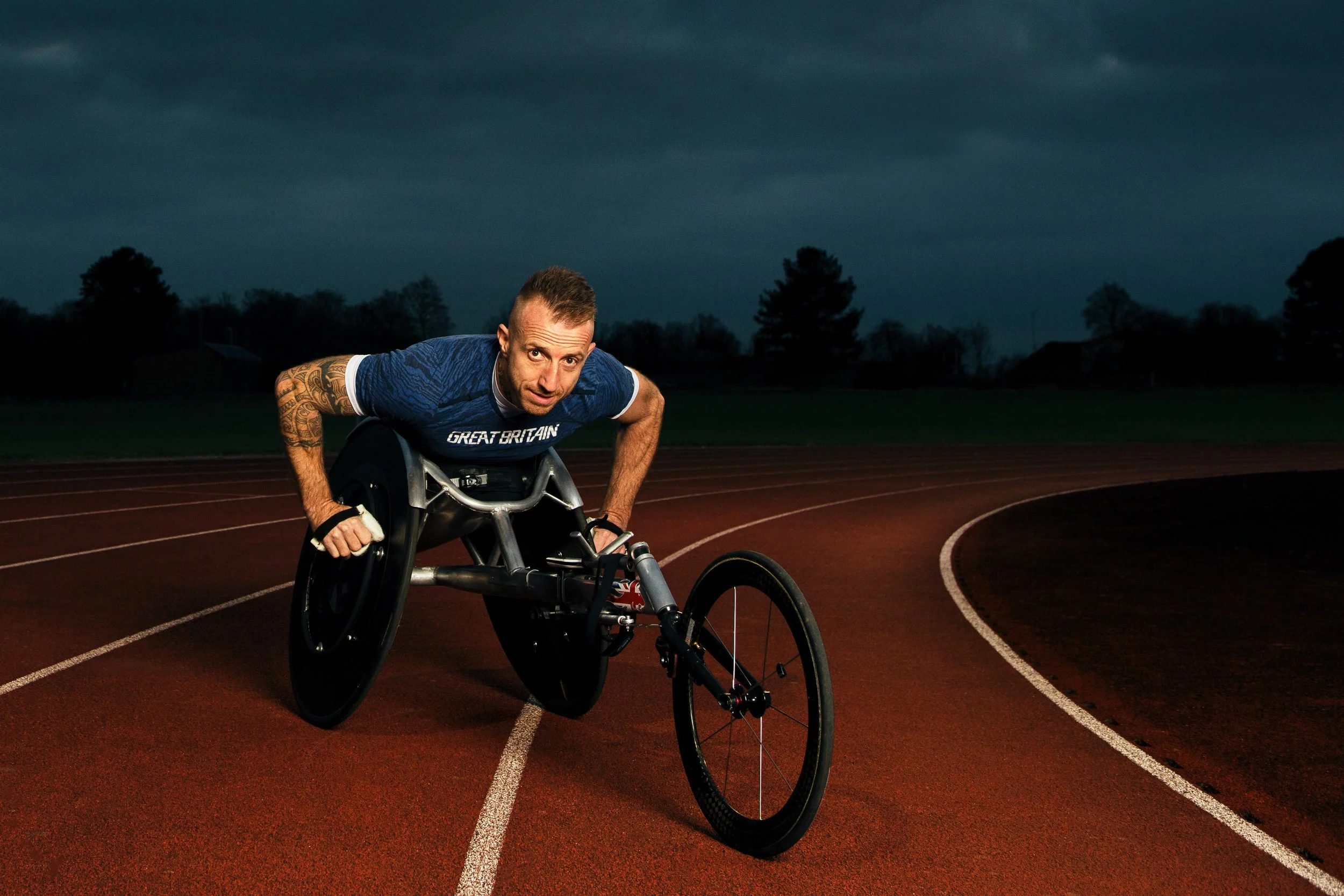 A male athlete in a wheelchair racing on an outdoor track during sunset or dusk, wearing a blue shirt that says 'Great Britain'.