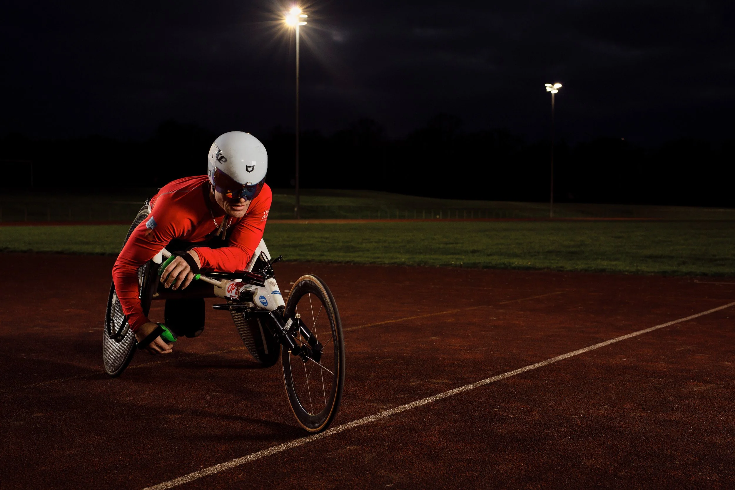 A male cyclist wearing a helmet and sunglasses biking on a track at night under bright floodlights.