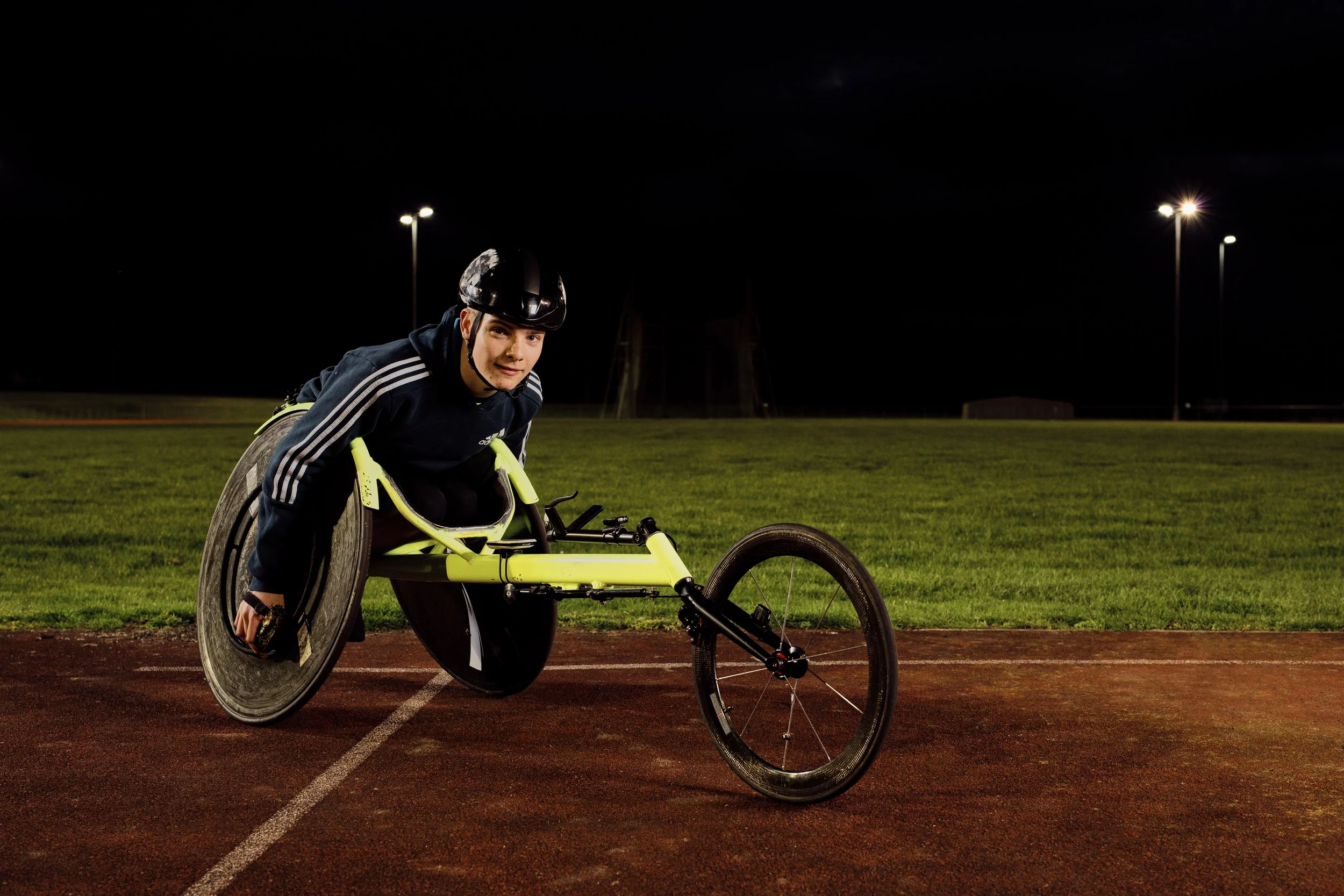 A male athlete in a wheelchair on a running track at night, wearing a helmet, preparing for a race.