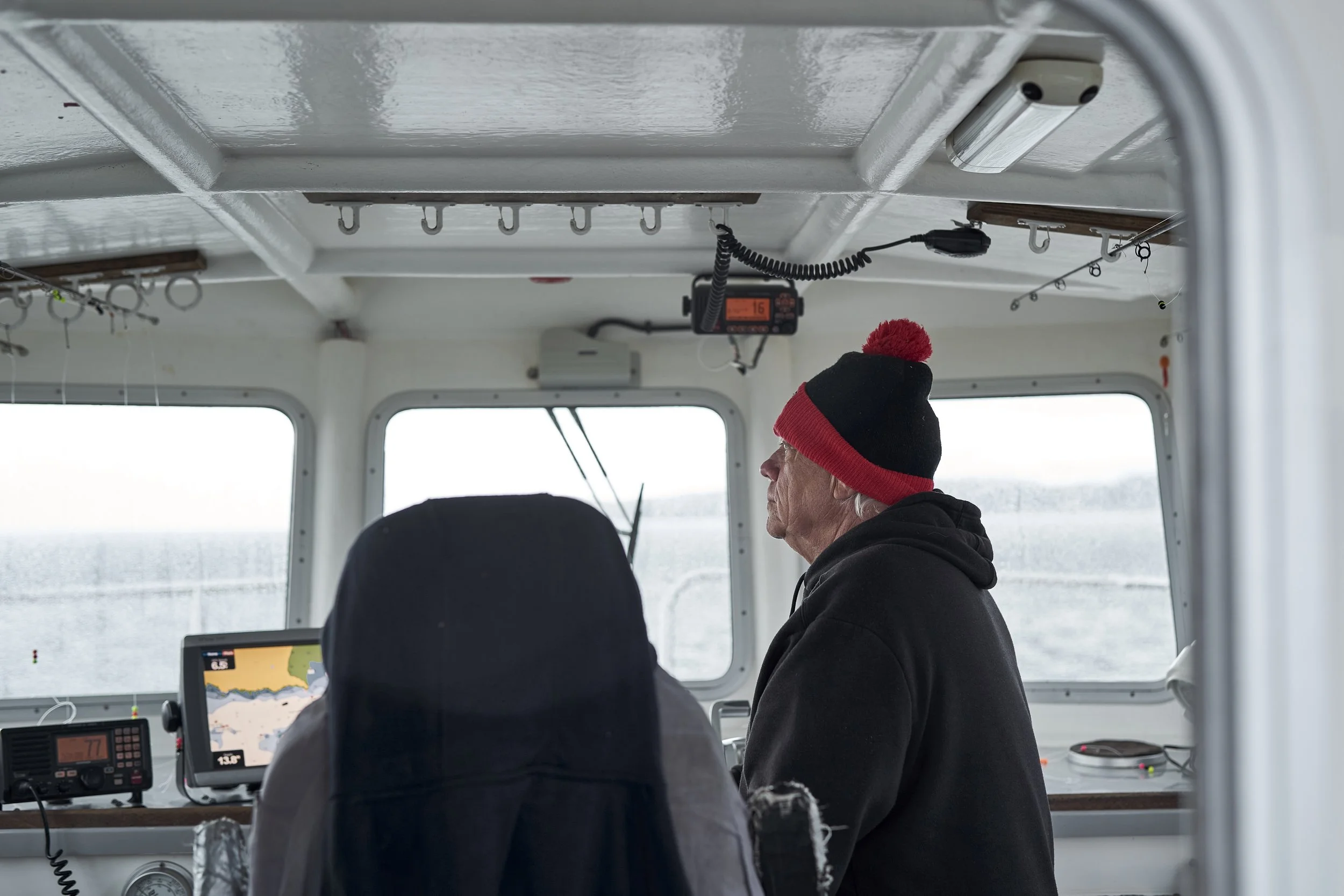 An older man wearing a black and red beanie and black hoodie sitting in a boat's control room, looking out the window at the water.