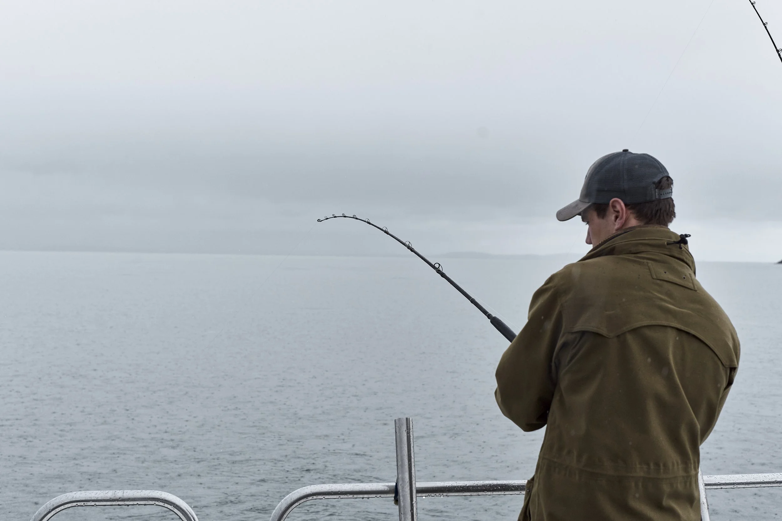 Man fishing off a boat in a gray, overcast sky and calm water.
