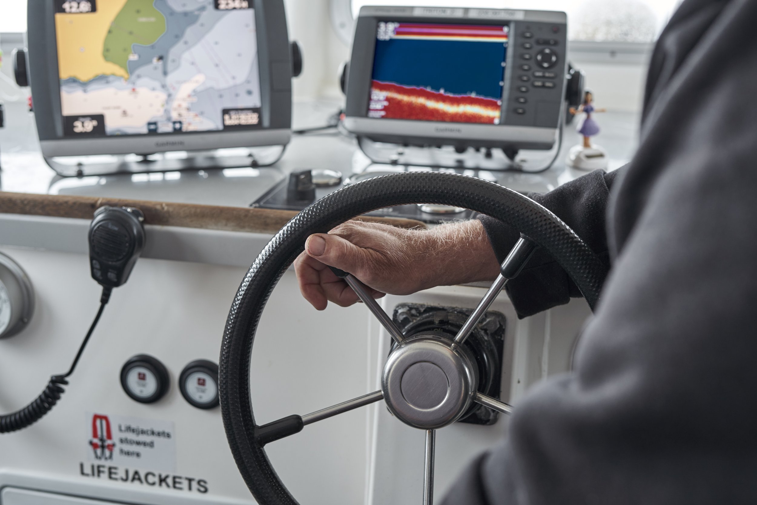 Person operating a boat with navigation and sonar screens in the background, a walkie-talkie hanging on the dashboard, and a sticker reading "LIFEJACKETS saved here."