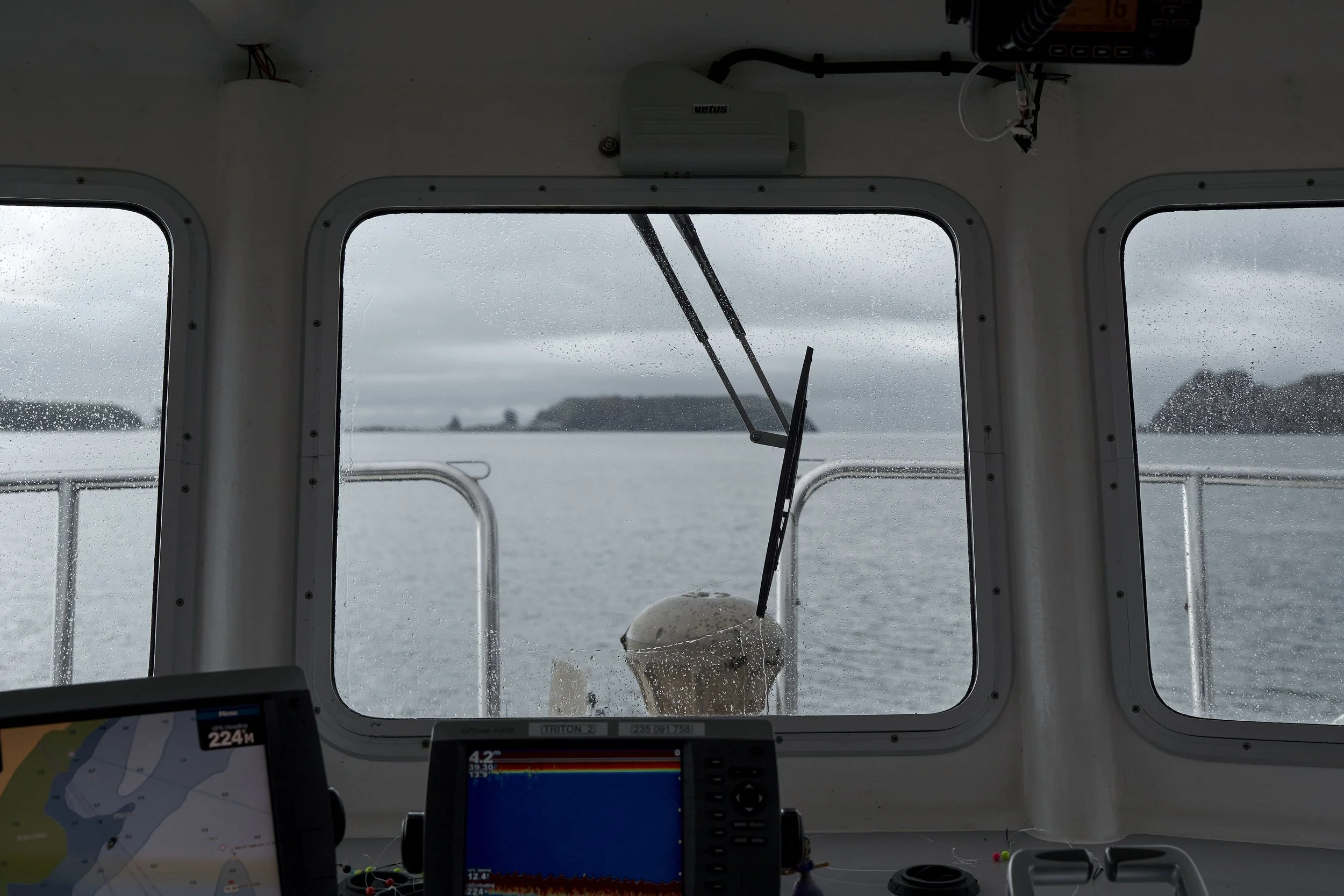 View through a boat's window showing water and distant islands on a rainy day, with boat controls and navigation screens visible inside the cabin.