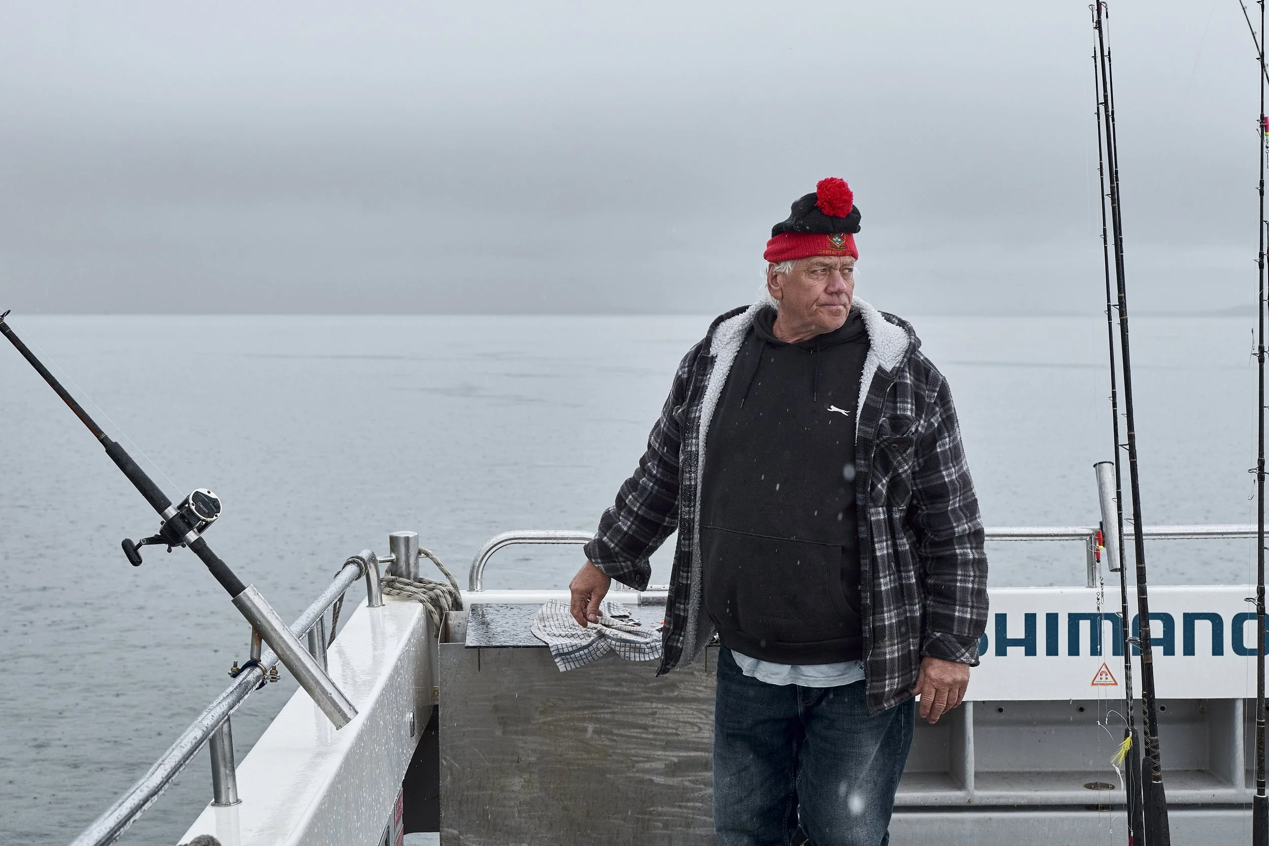 An older man in a black hoodie, plaid jacket, and red and black beanie stands on a fishing boat with fishing rods, looking out over the gray, calm water and sky.
