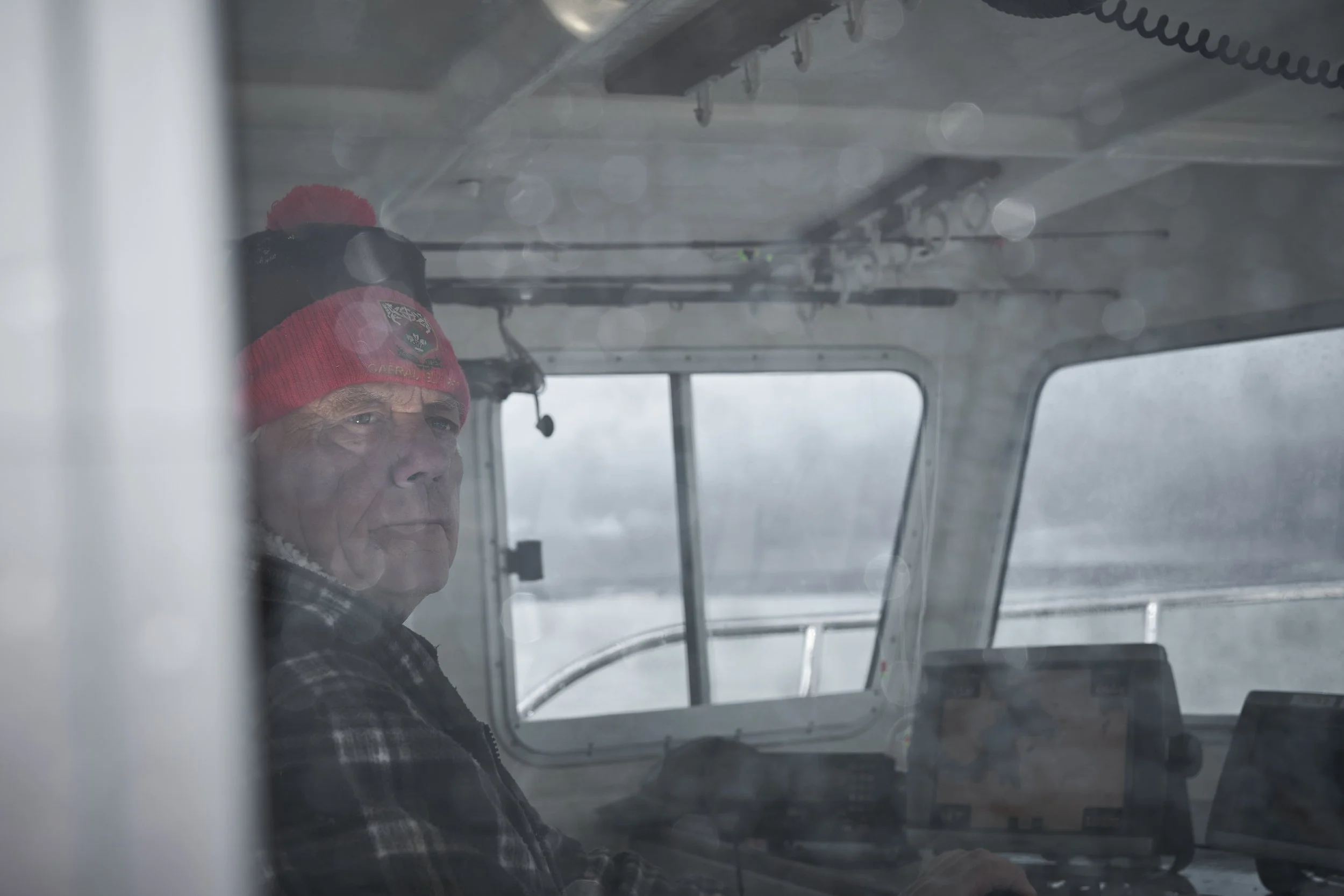 A man with a serious expression wearing a red beanie with a pom-pom and a plaid shirt, sitting inside a boat cockpit with snow outside the window.
