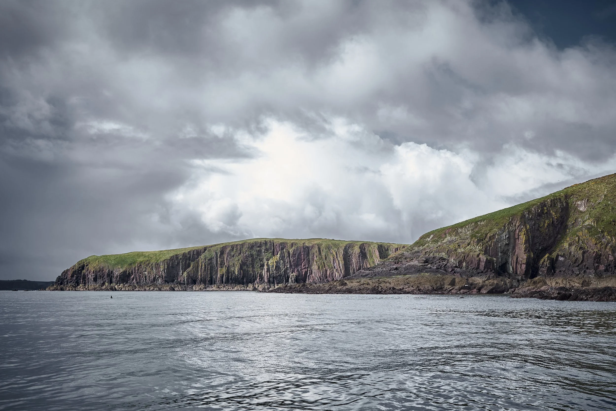 Scenic view of a rocky coastline with green grassy tops, under a cloudy sky, reflecting off the water.