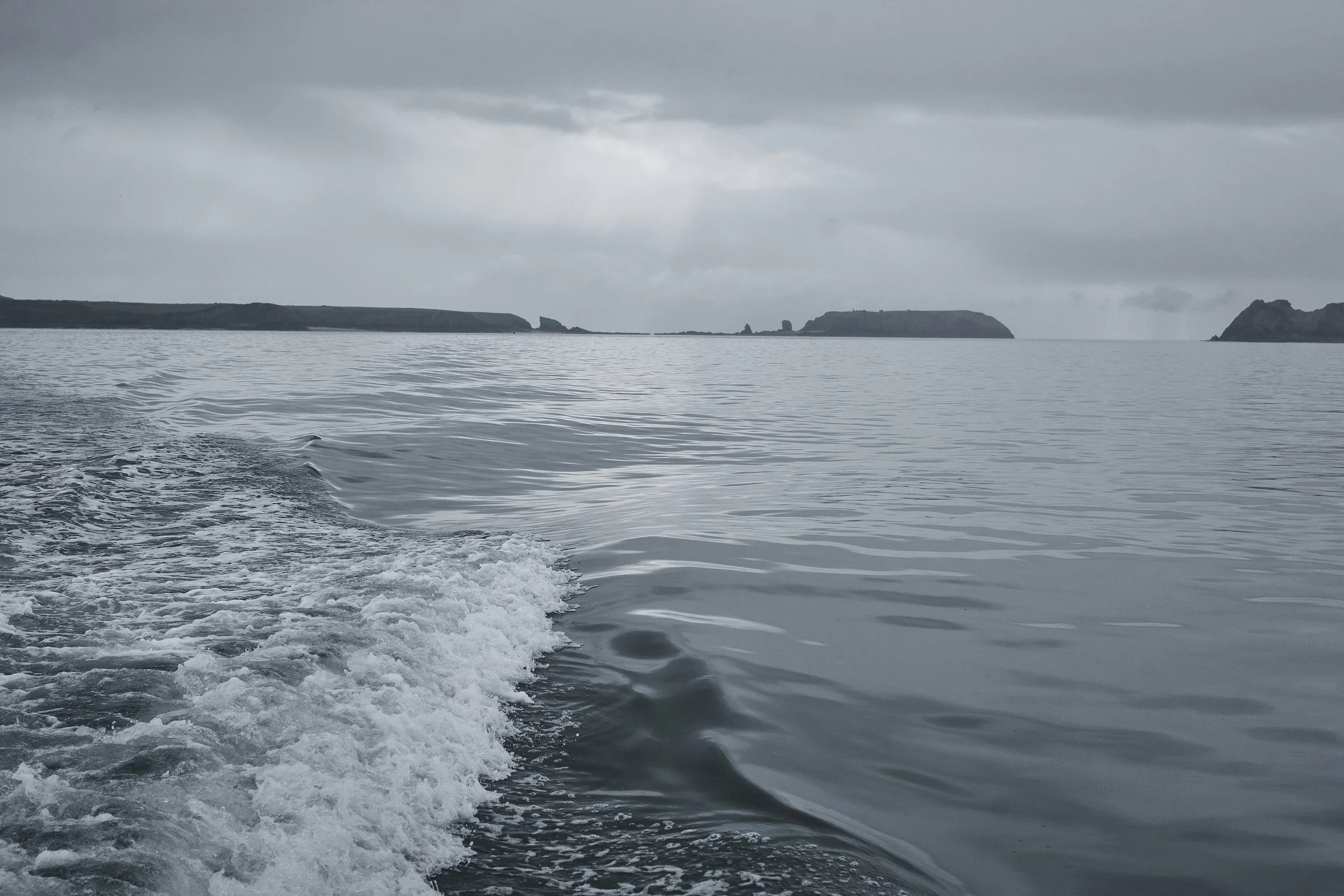 View of a calm ocean with a boat's wake in the foreground, and distant landforms and cliffs under a cloudy sky.