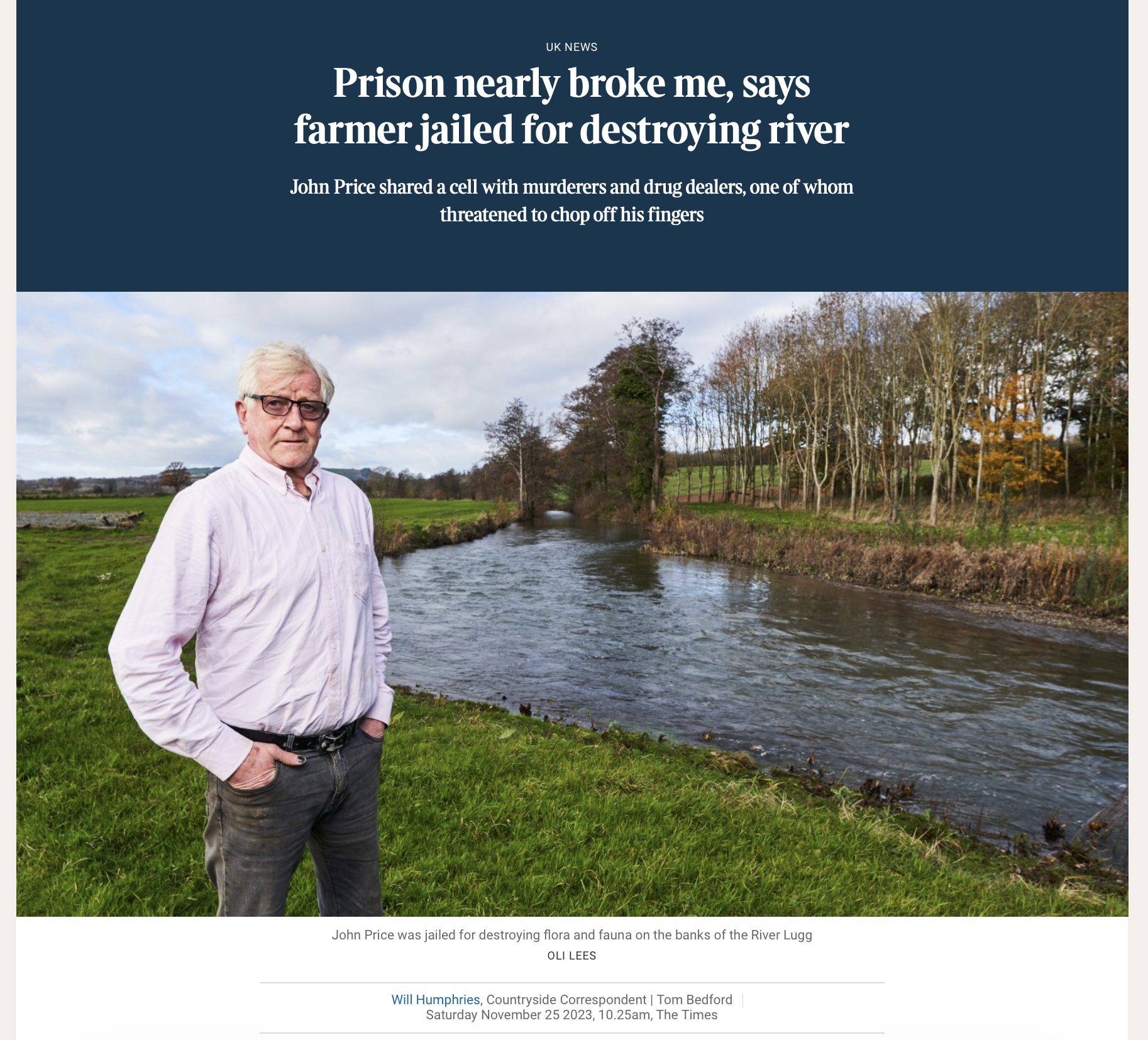 A man in a light pink shirt and glasses standing by a river with trees and green fields in the background.