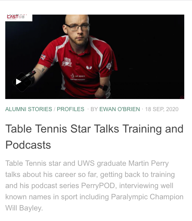 A man with glasses and a beard wearing a red and white sports shirt, sitting at a table with a black background.