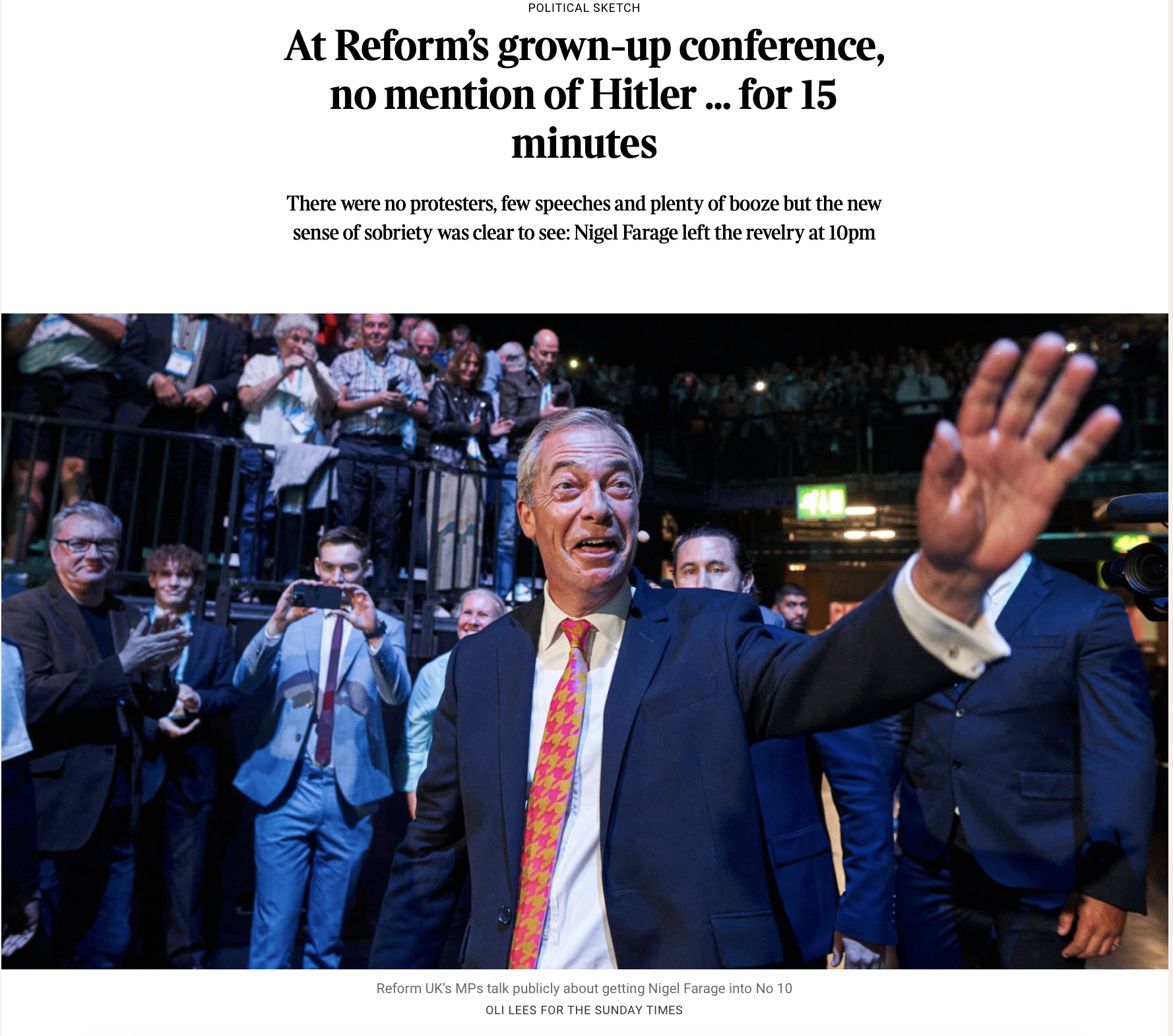 Nigel Farage, a man in a suit with a colorful tie, waving to a crowd at a political event. Behind him, there are spectators and supporters, some taking photos and videos. The setting appears to be an indoor or enclosed venue. The caption notes this i