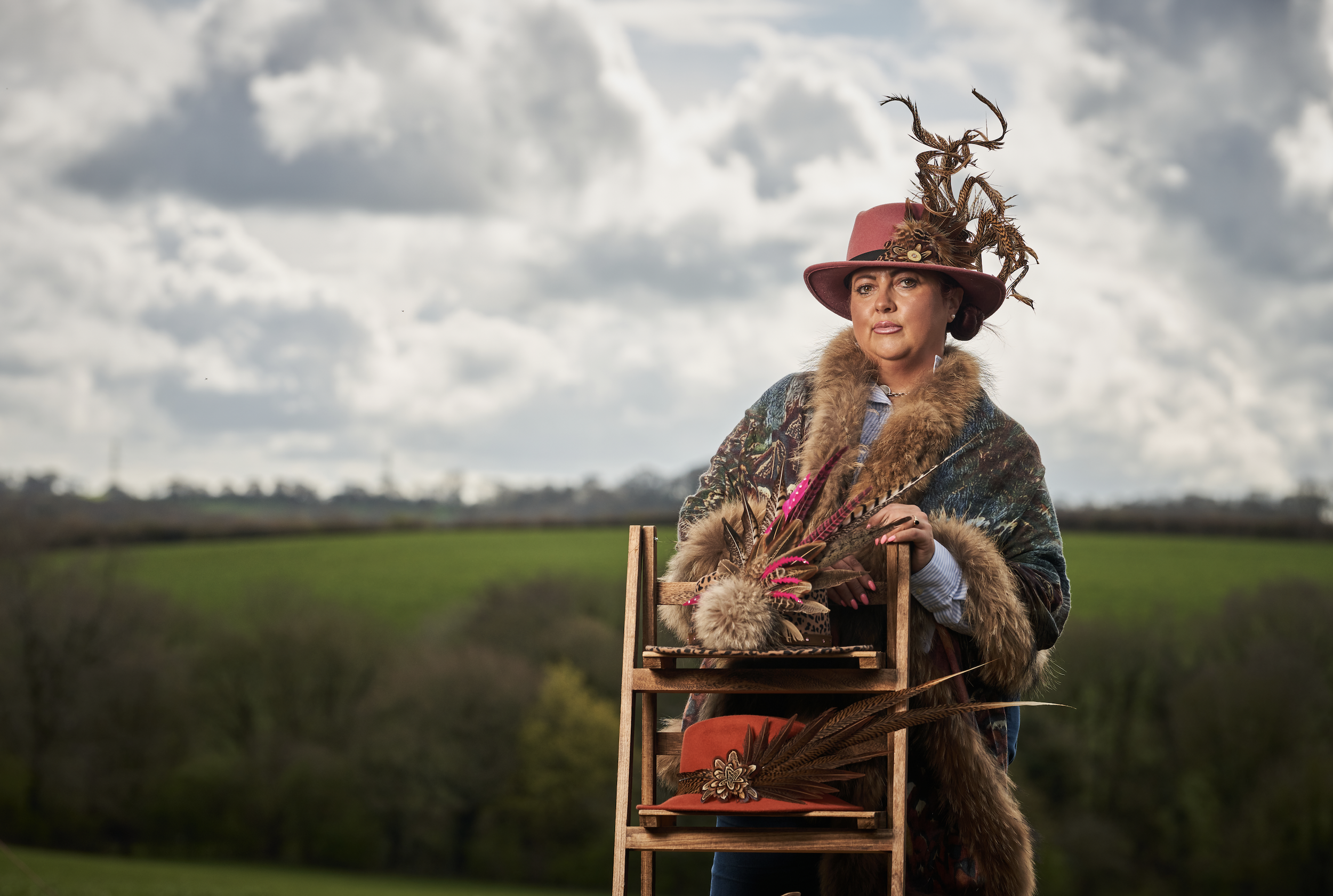 A woman outdoors wearing a pink hat with feathers and a fur-lined coat, standing behind a wooden display rack with multiple hats and feather accessories, against a cloudy sky and green landscape in the background.