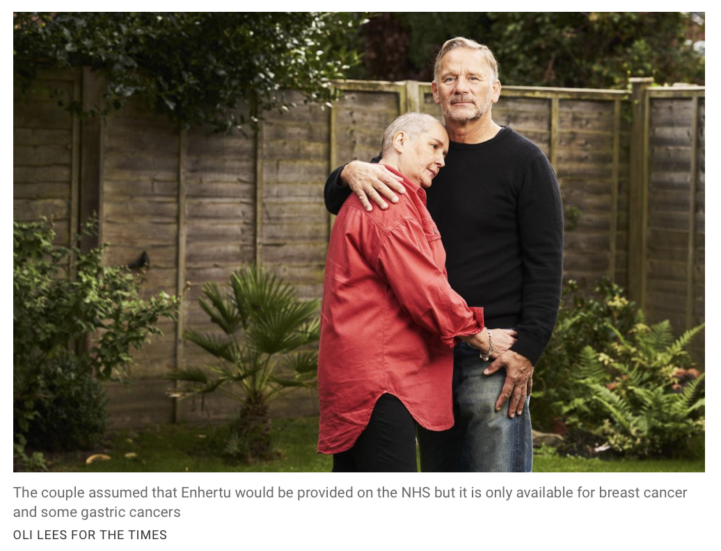 A man and a woman stand closely together in a garden, with the woman resting her head on the man's chest. The man is wearing a black sweater, and the woman is in a red jacket. They are embracing, and the background features a wooden fence and greener