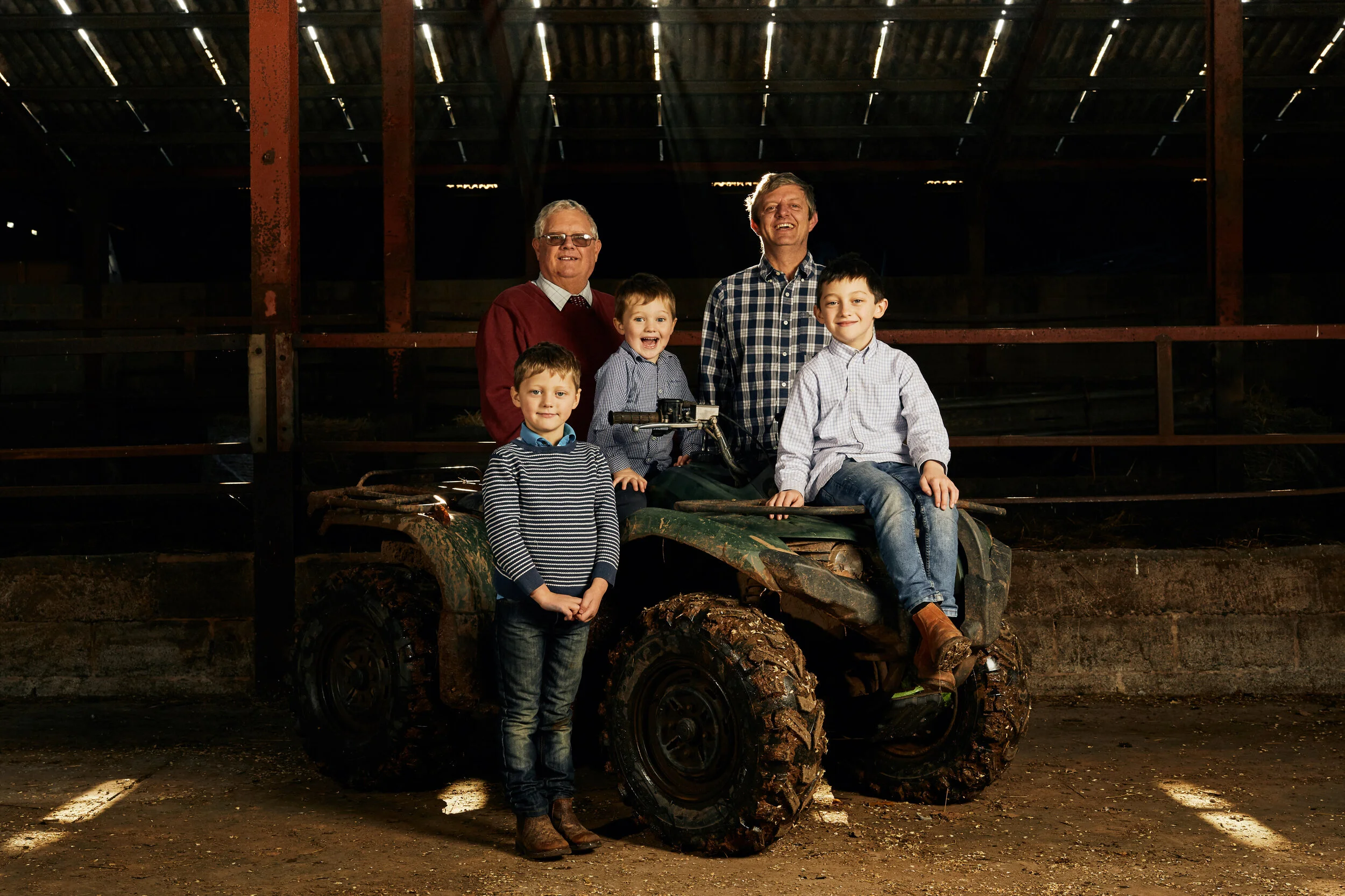 Group of five people, including three boys and two men, in a barn with dirt floor, standing around a muddy ATV, with barn structure and planks in the background.