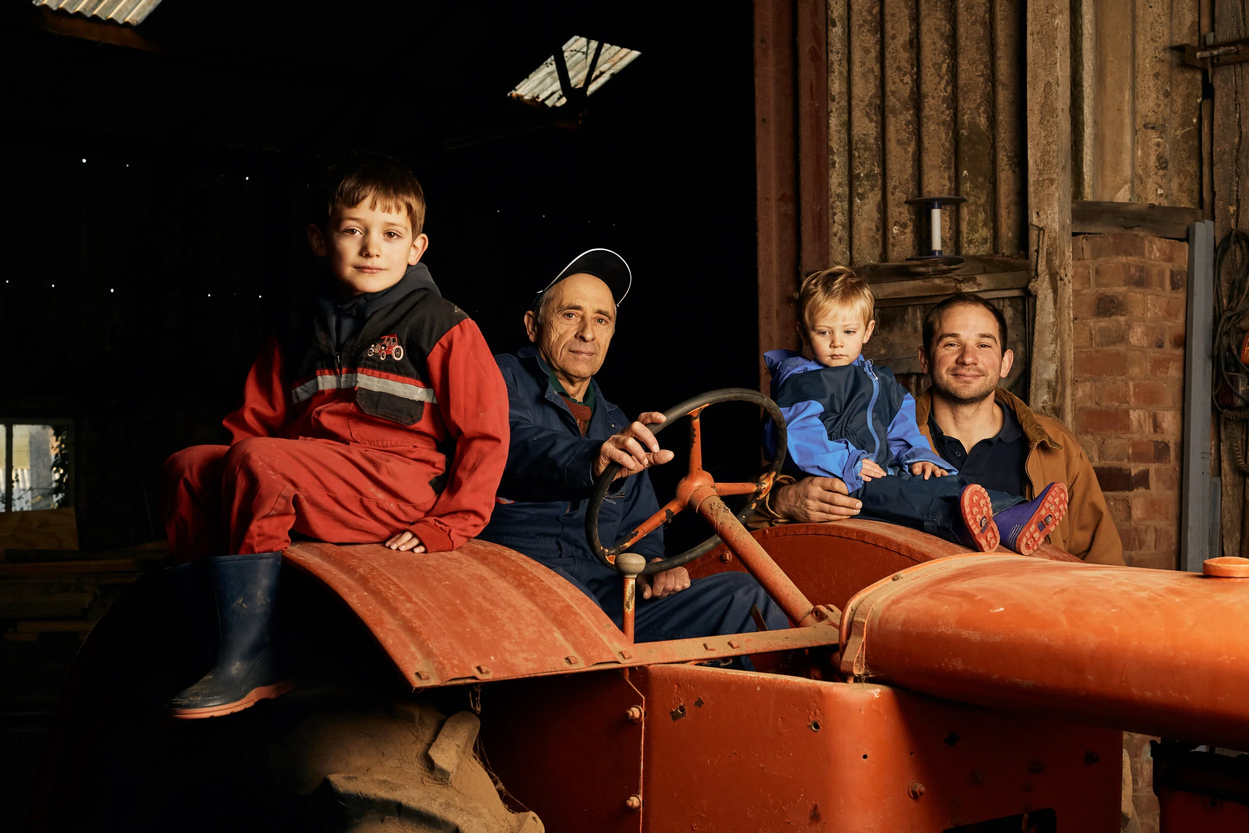 Four males sitting on a vintage red tractor inside a rustic barn. One young boy in red coveralls, an older man with a cap, a young girl in blue, and a smiling man in a brown jacket.