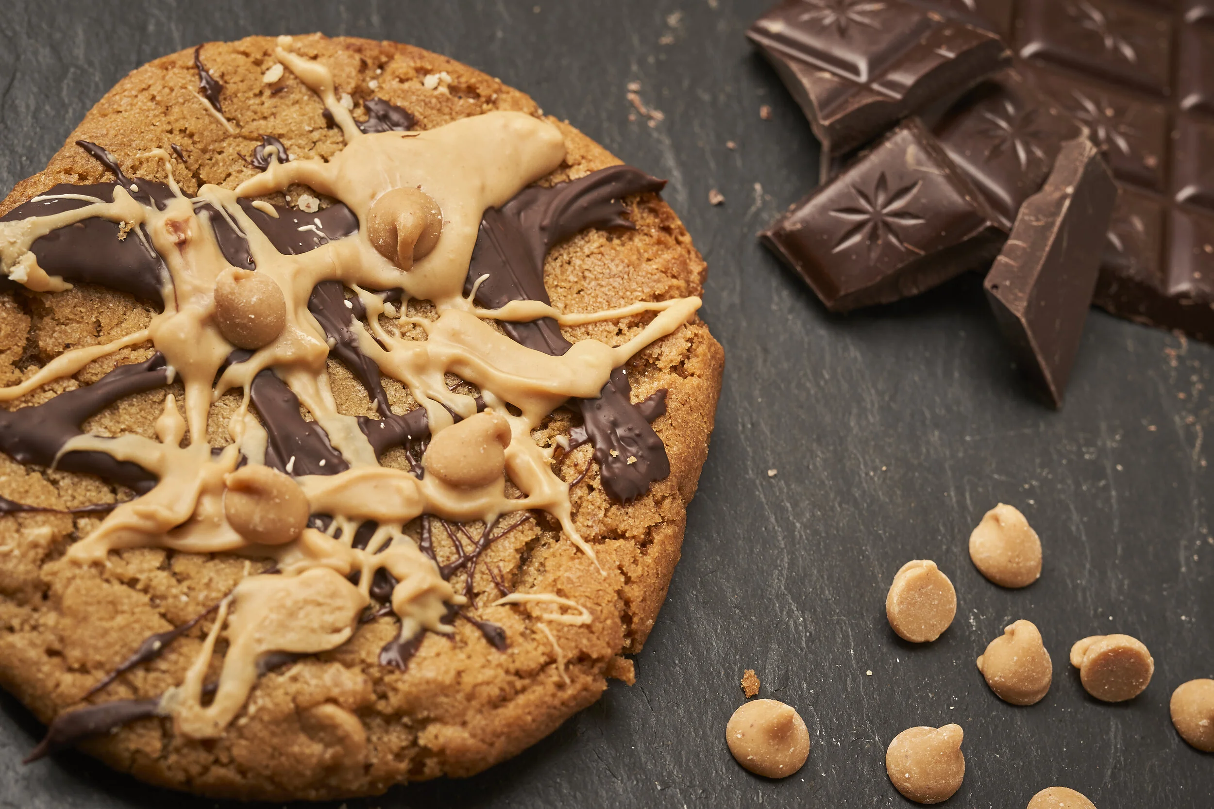Chocolate chip cookie decorated with drizzles of milk and dark chocolate and small round candies, with broken chocolate pieces nearby on a dark surface.
