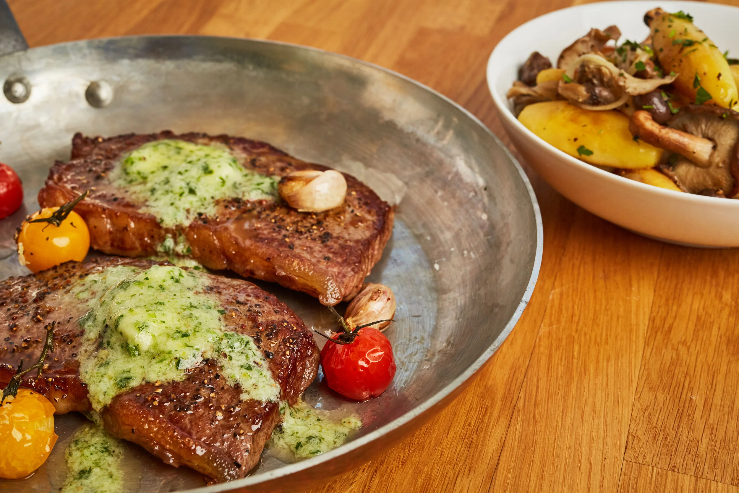 Two cooked steaks garnished with green herb butter, alongside cherry tomatoes and garlic cloves, on a metal serving dish. A bowl of roasted potatoes with mushrooms is visible in the background.
