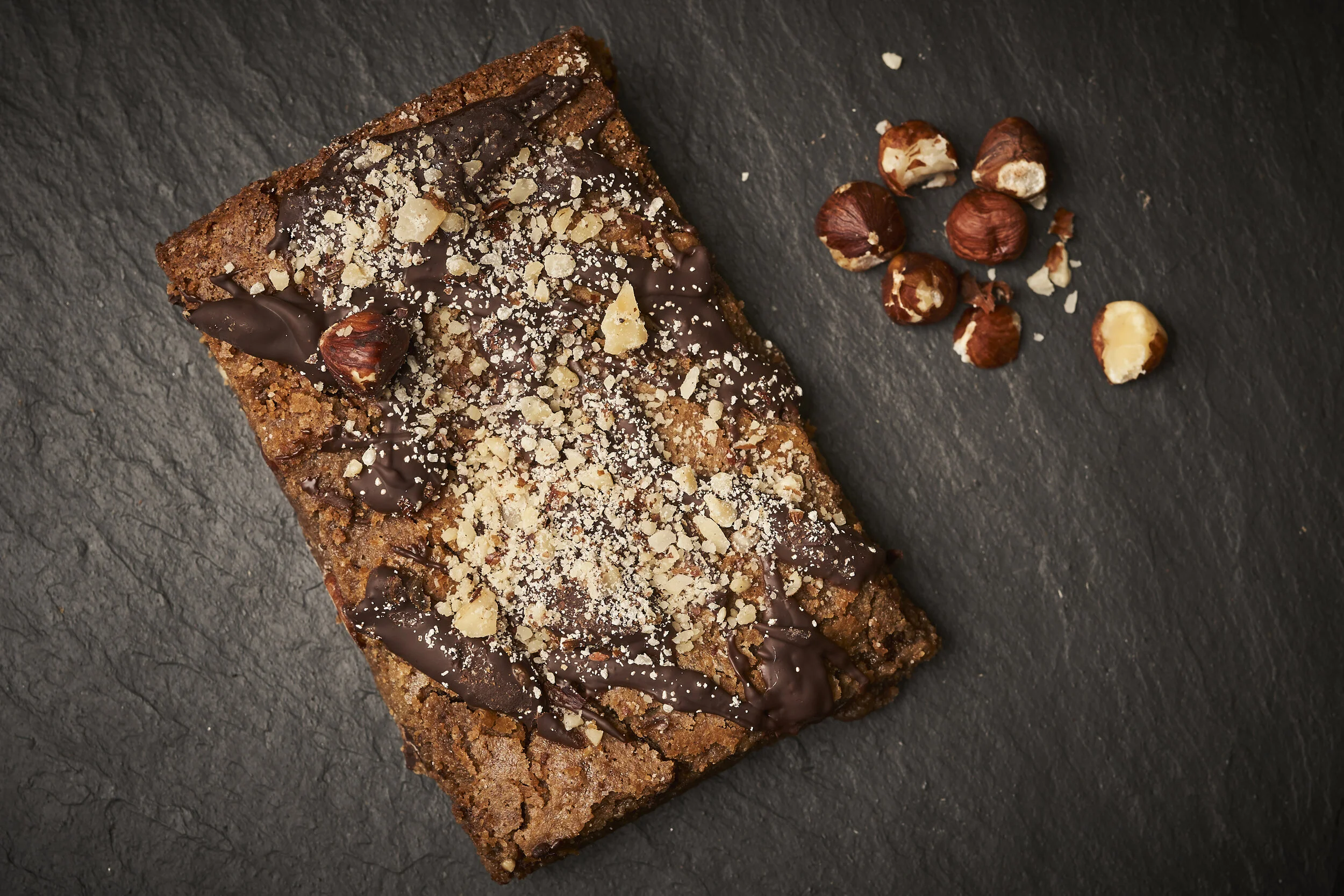 Chocolate brownie topped with chopped nuts and drizzled with chocolate, with hazelnuts scattered beside it on a black slate surface.