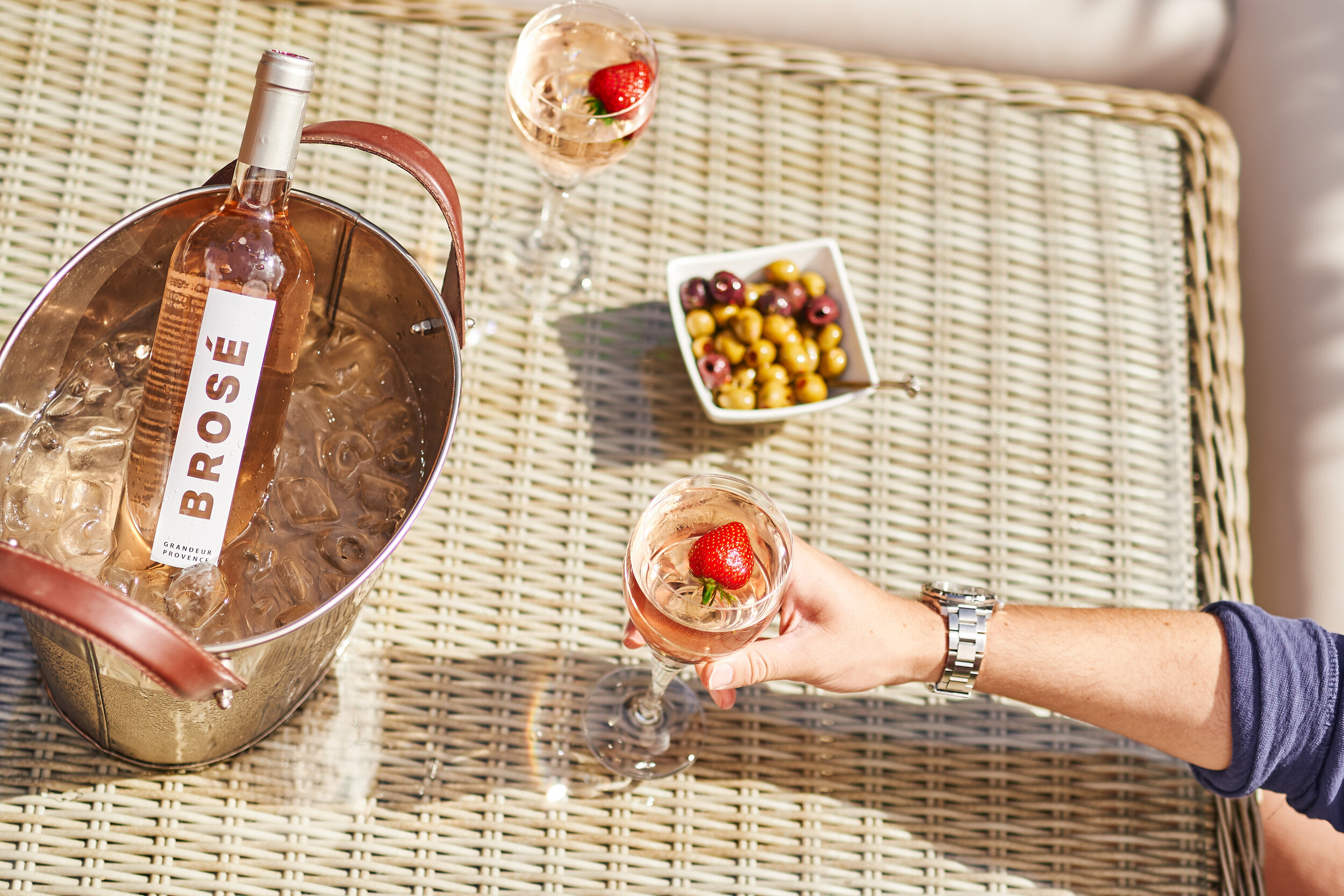 A person holding a glass of rosé wine with a strawberry garnish on a table with other glasses, a bottle of rosé wine in an ice bucket, a bowl of mixed olives, and a small bowl with additional strawberries and grapes.
