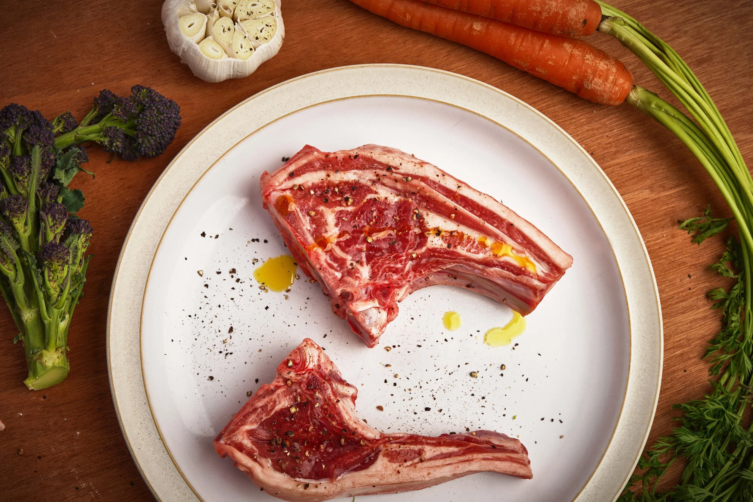 Raw lamb chops seasoned with black pepper, on a white plate, with vegetables including carrots, garlic, broccoli, and herbs around the plate.