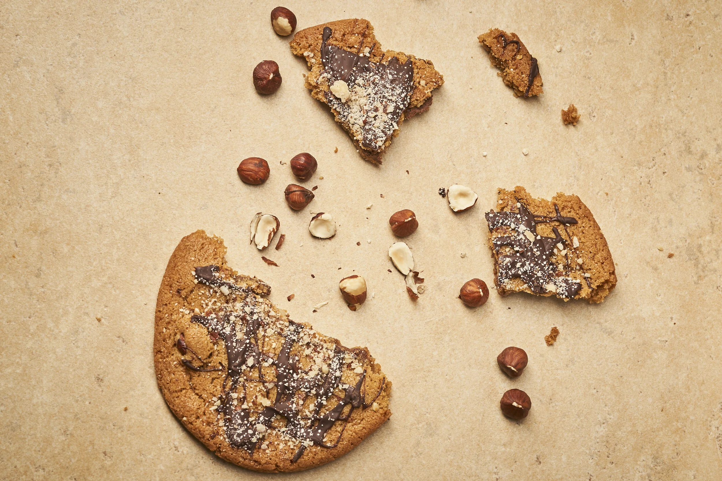 Broken chocolate chip cookie with hazelnuts and scattered hazelnuts on parchment paper.