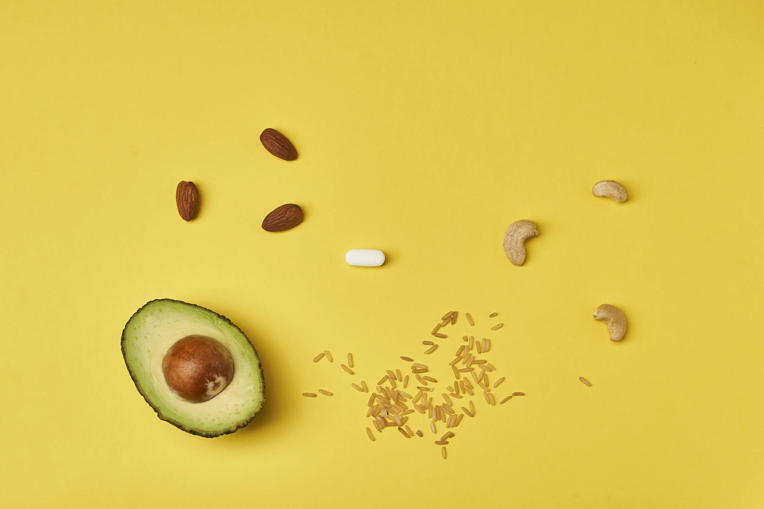 Half an avocado with pit, scattered almonds, cashews, rice grains, and one white pill on a yellow background.