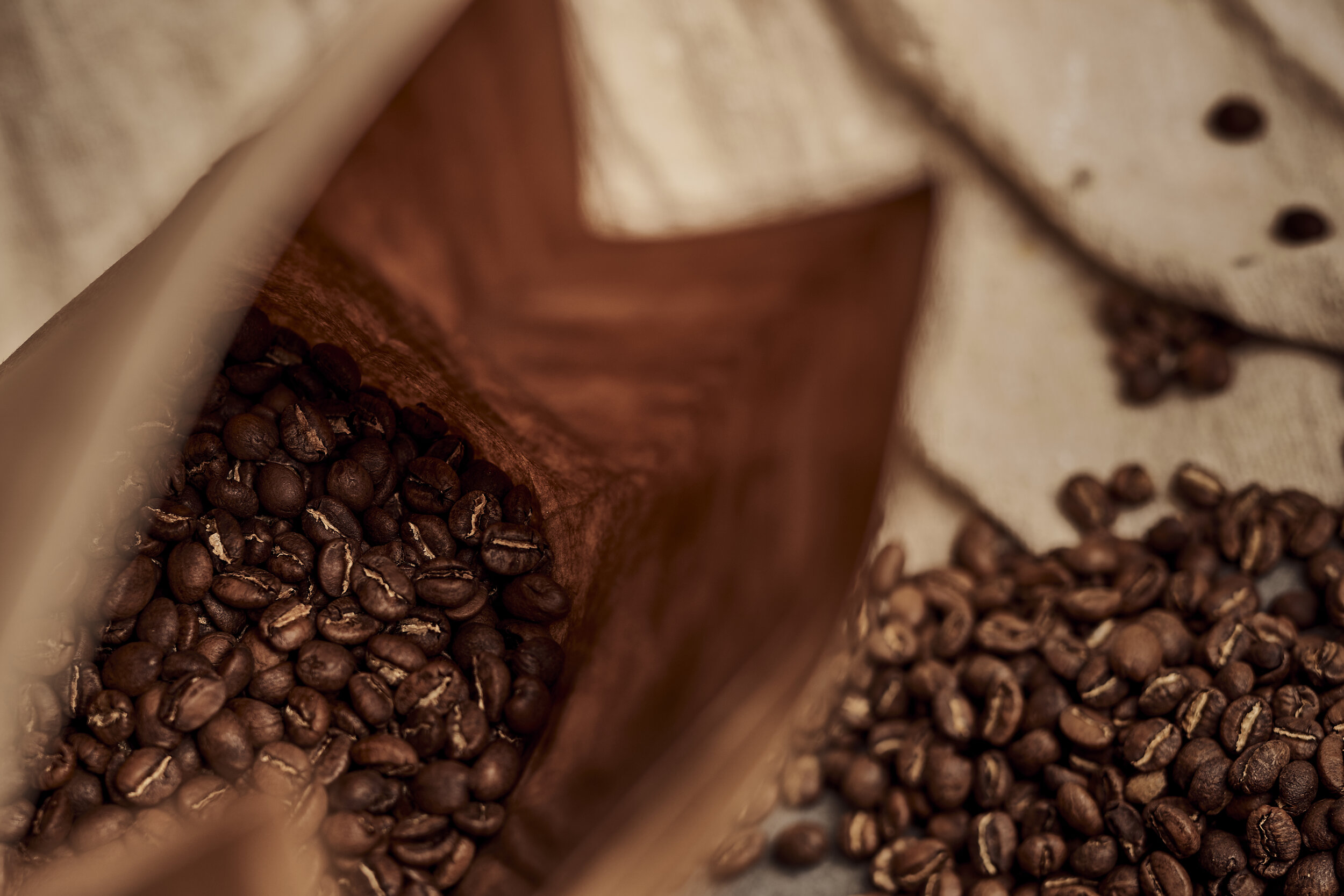 Close-up of roasted coffee beans in and around an open paper bag on a cloth surface.