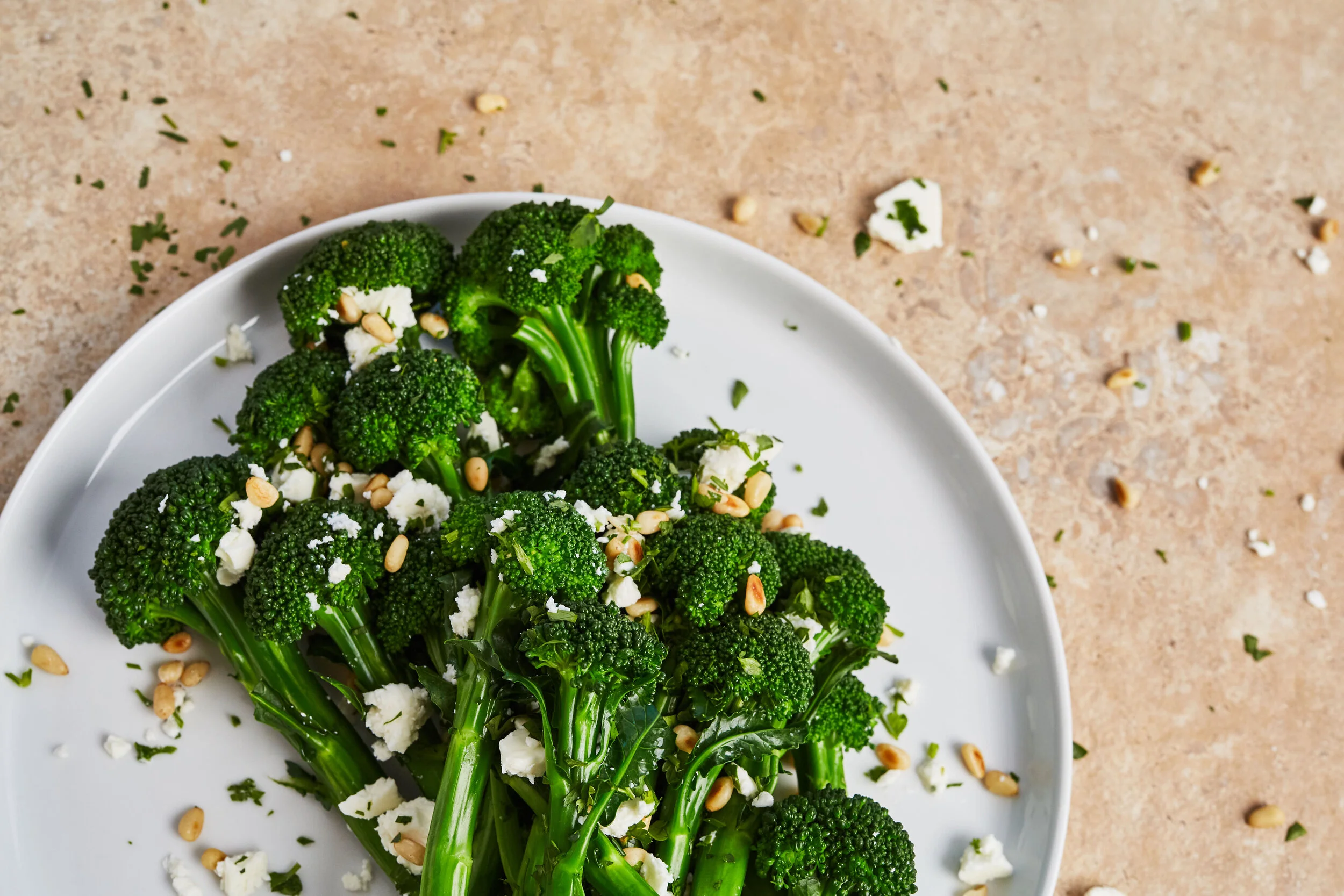 Fresh broccoli florets garnished with crumbled cheese and pine nuts on a white plate, with scattered herbs and nuts on a beige surface.