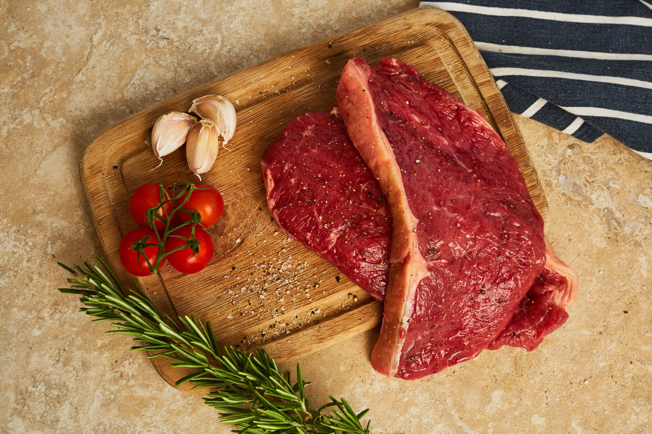 Raw beef steak with garlic cloves, cherry tomatoes on vine, and rosemary on a wooden cutting board.