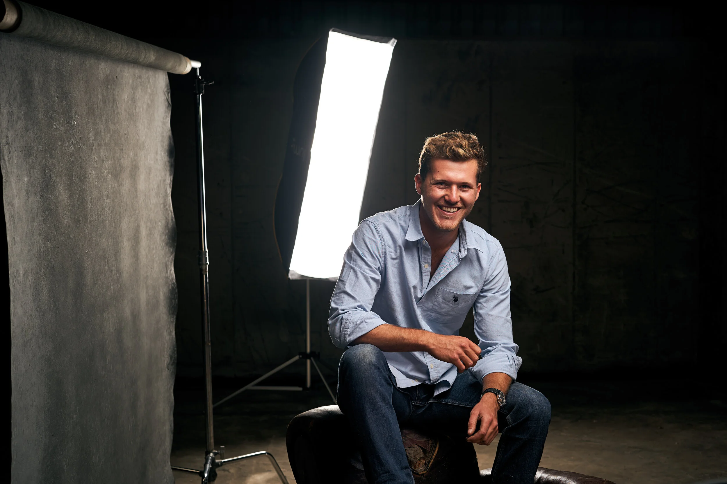 Young man in light blue shirt smiling while sitting on a stool in front of studio lighting and a backdrop.
