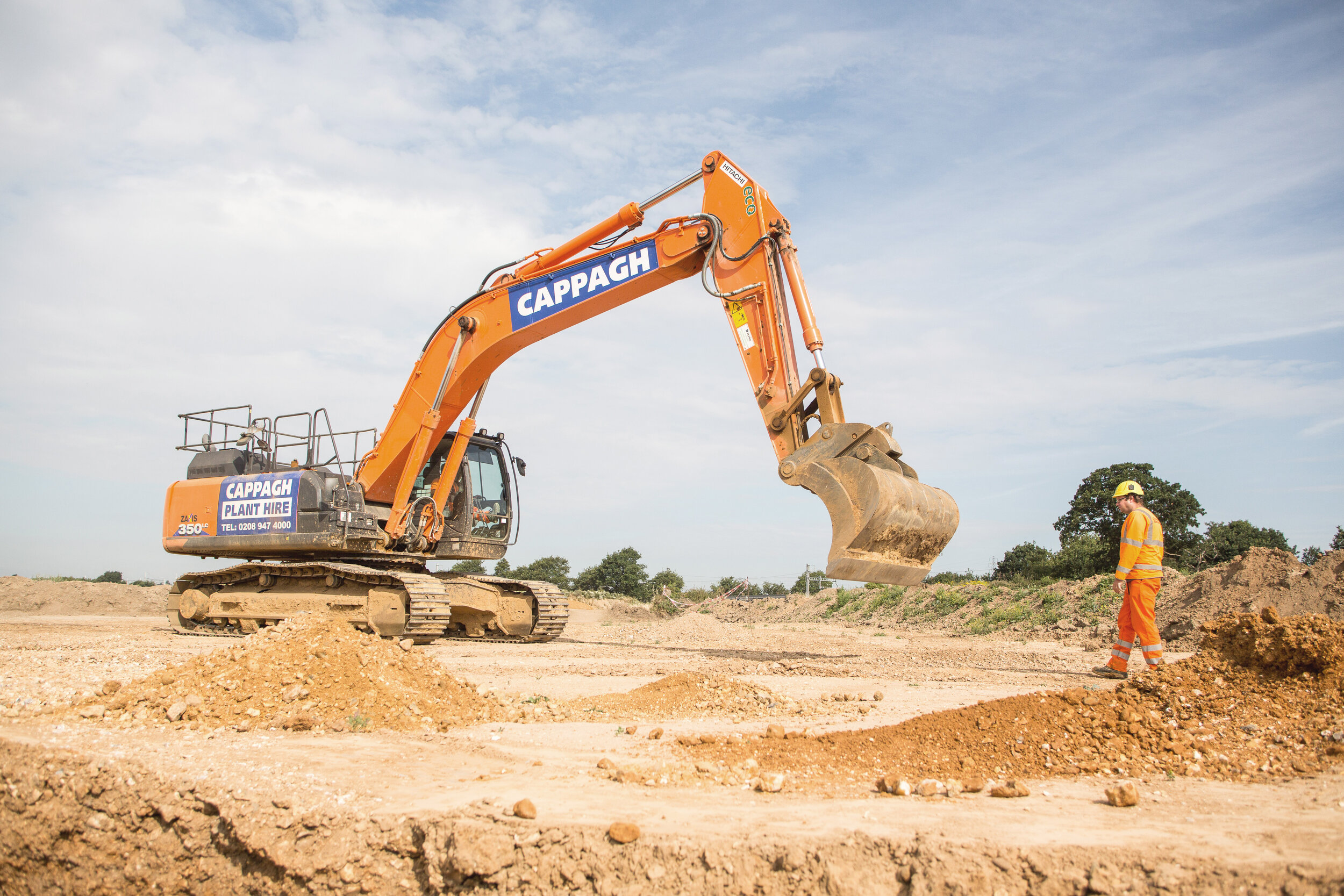 Construction site with an orange excavator and a worker in orange safety gear and a yellow helmet.