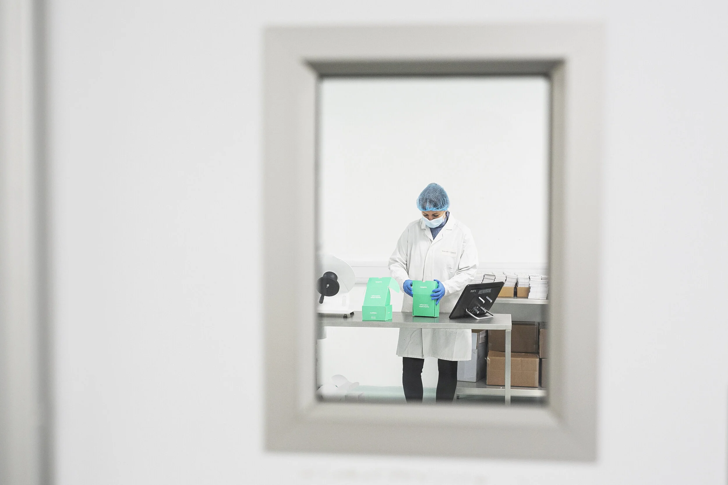 A lab technician in protective gear working at a table, viewed through a small window.