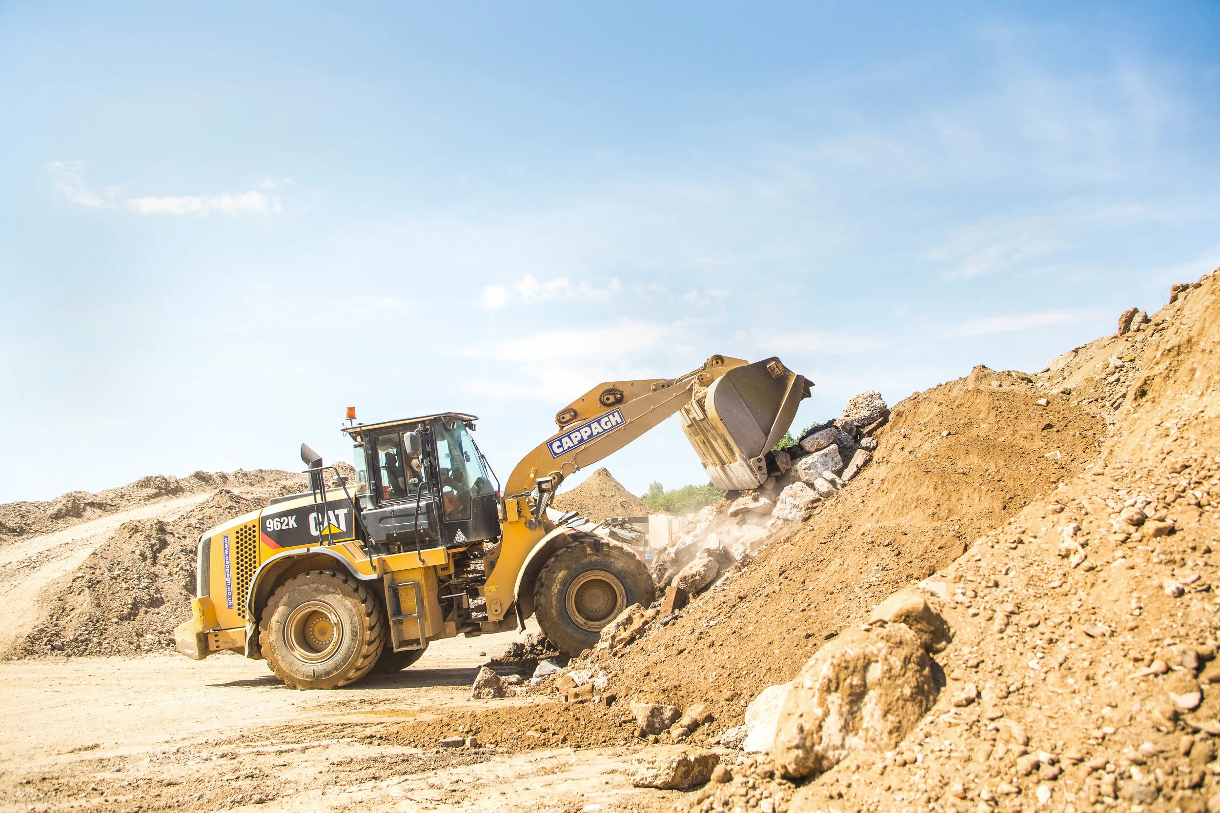 A yellow Caterpillar excavator loading rocks and dirt on a construction site under a clear blue sky.