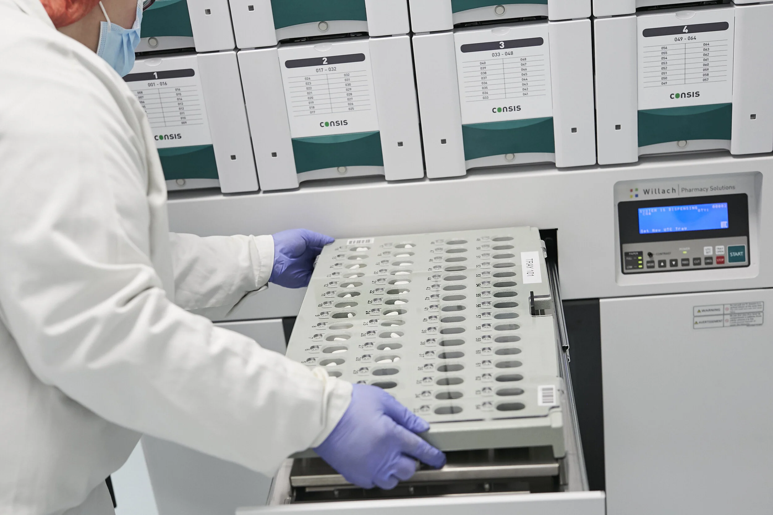 A healthcare worker wearing a face mask, gloves, and a white lab coat handling a tray of test tubes in a laboratory with medical storage boxes on the shelves.
