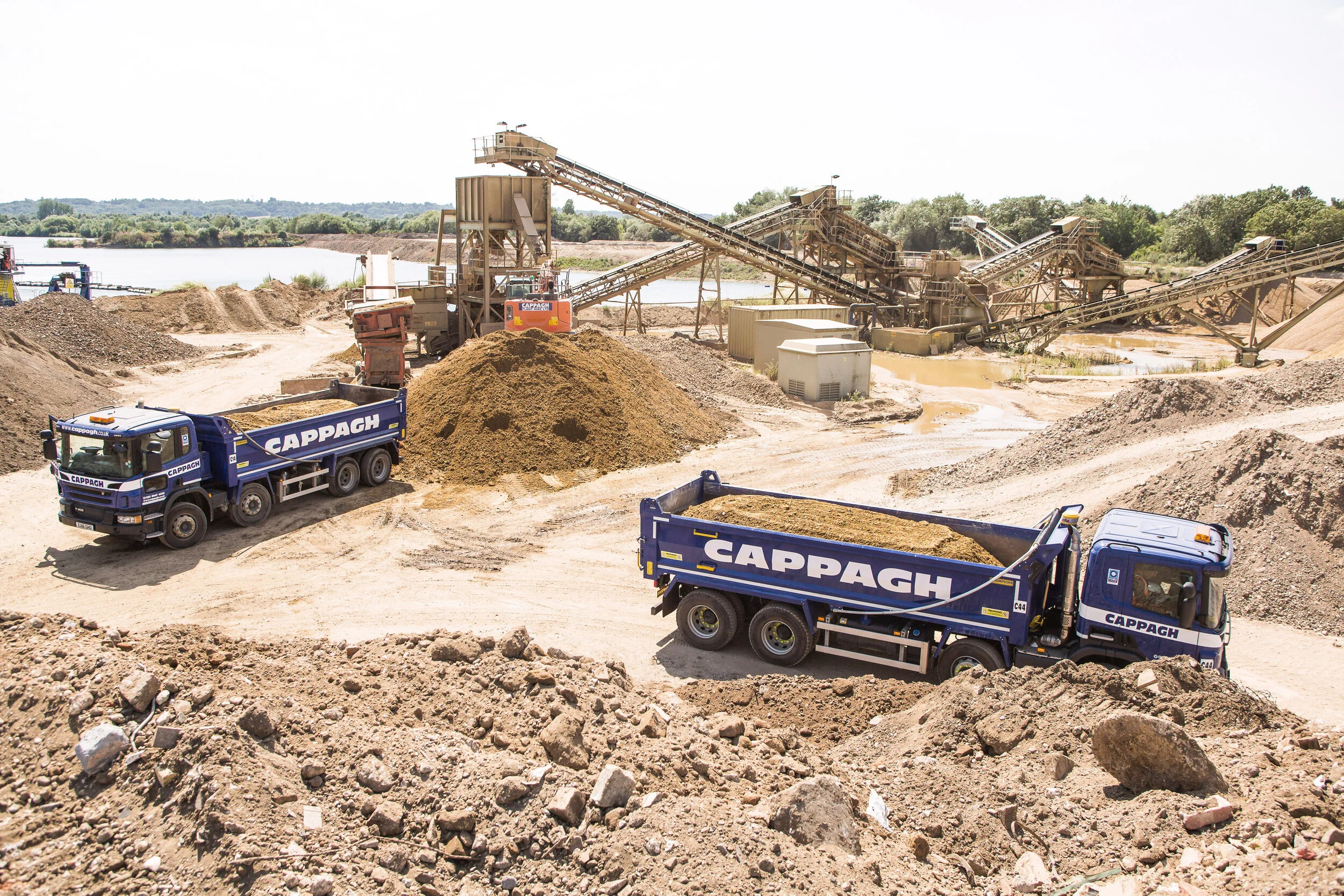 Construction site with two blue dump trucks labeled 'CAPPAGH' on a dirt road, piles of dirt, and explosives conveyor belts in the background.