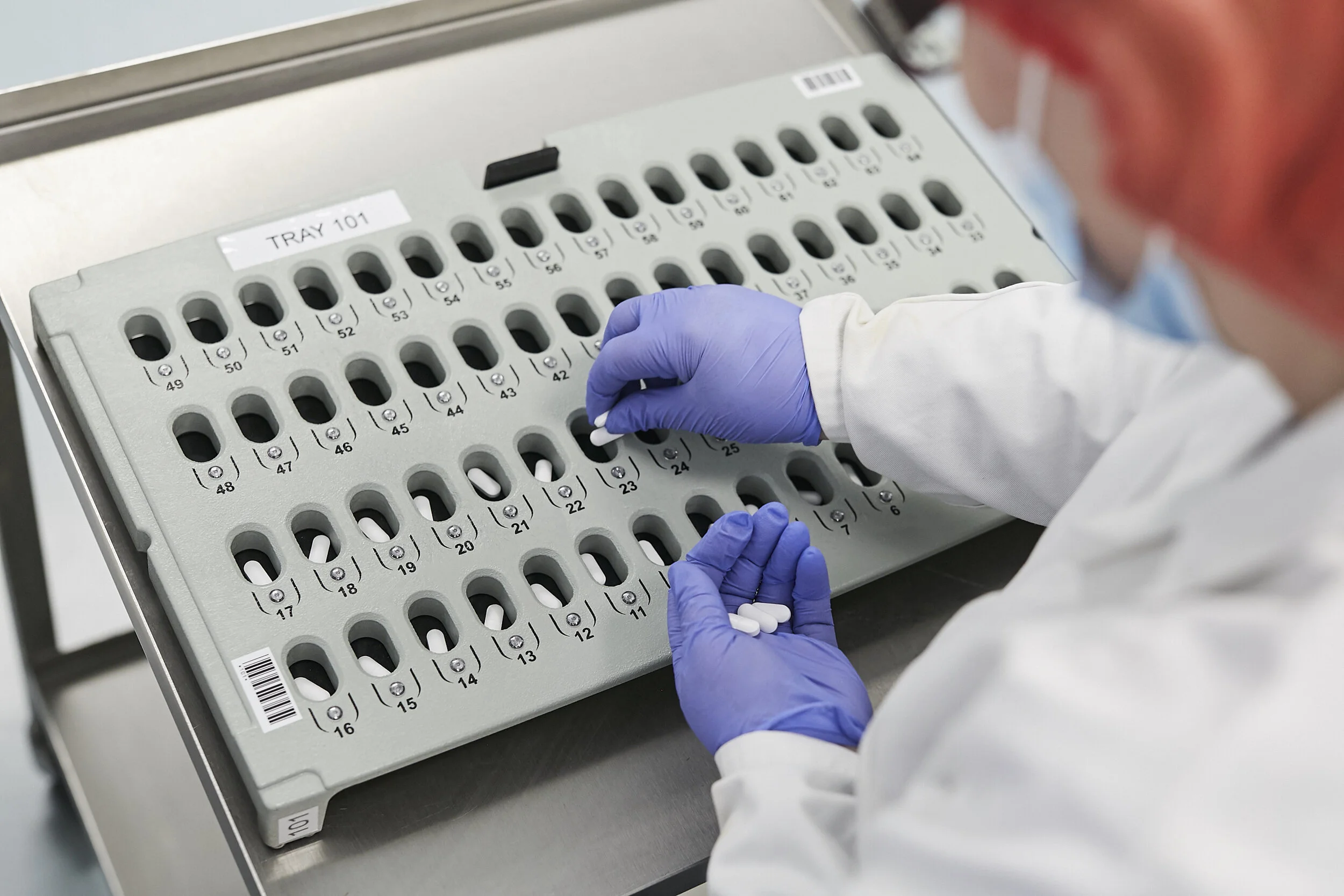 A healthcare worker wearing gloves and a mask handling pills from a numbered medication tray labeled 'TRAY 101'.