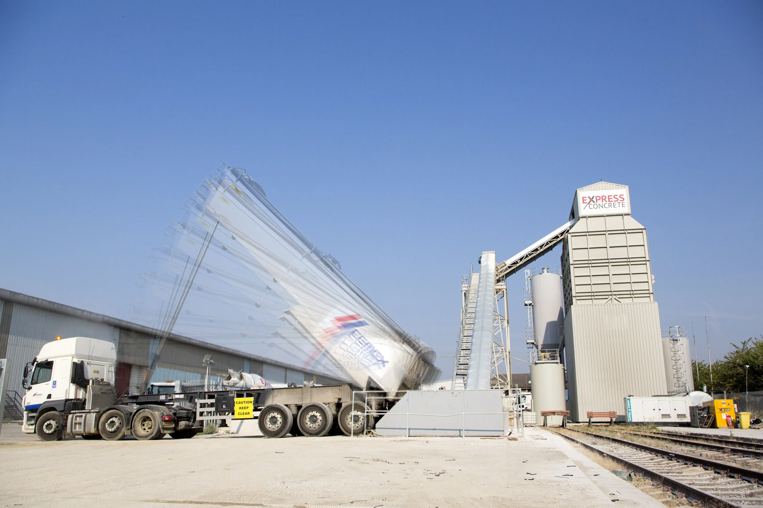 A cement truck is tipped over on its side with cement pouring out onto the ground at a cement plant, with a large industrial structure labeled EXPRESS CONCRETE and railway tracks nearby.
