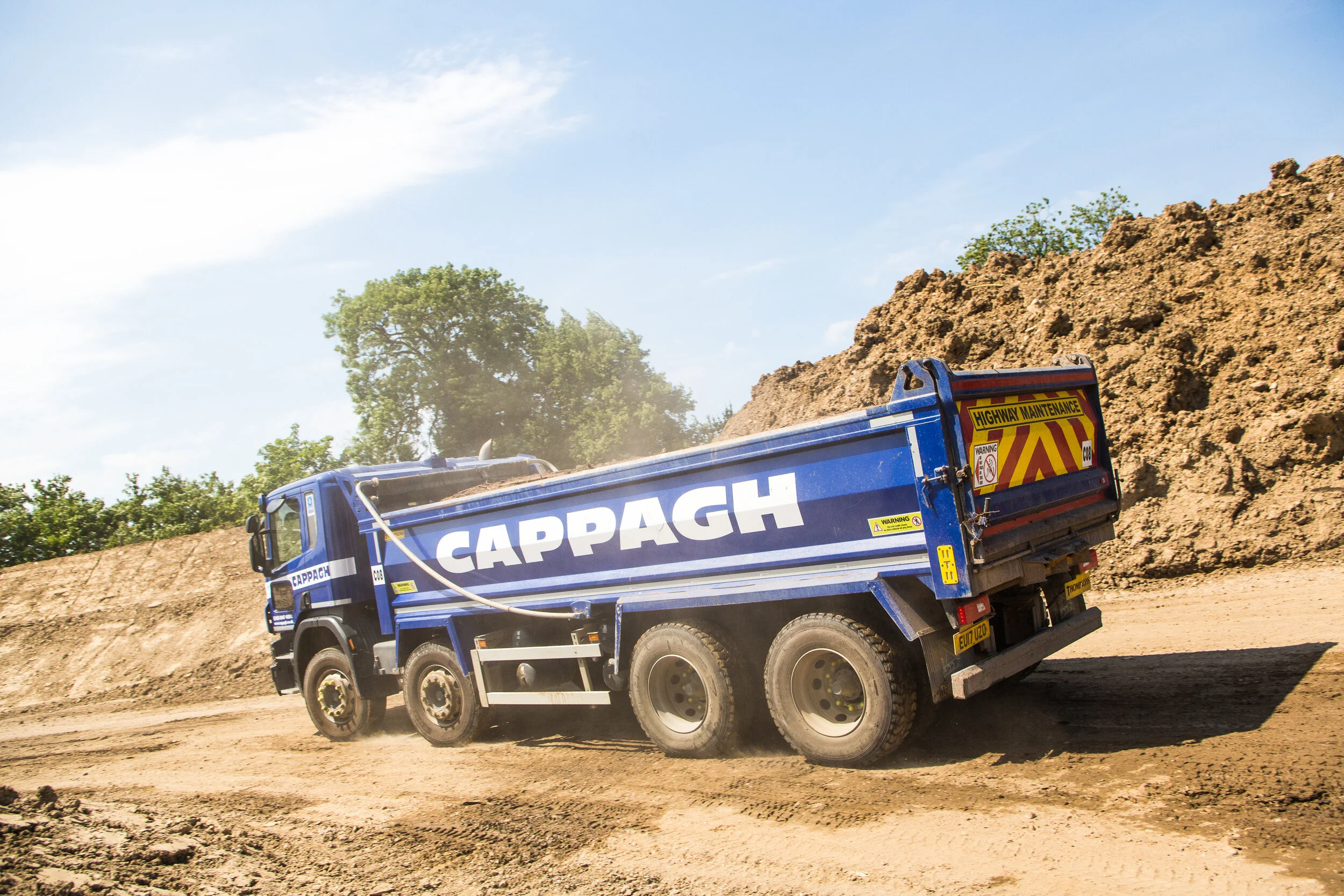 Blue Cappagh construction dump truck on dirt road at a construction site with dirt mounds and trees in the background.