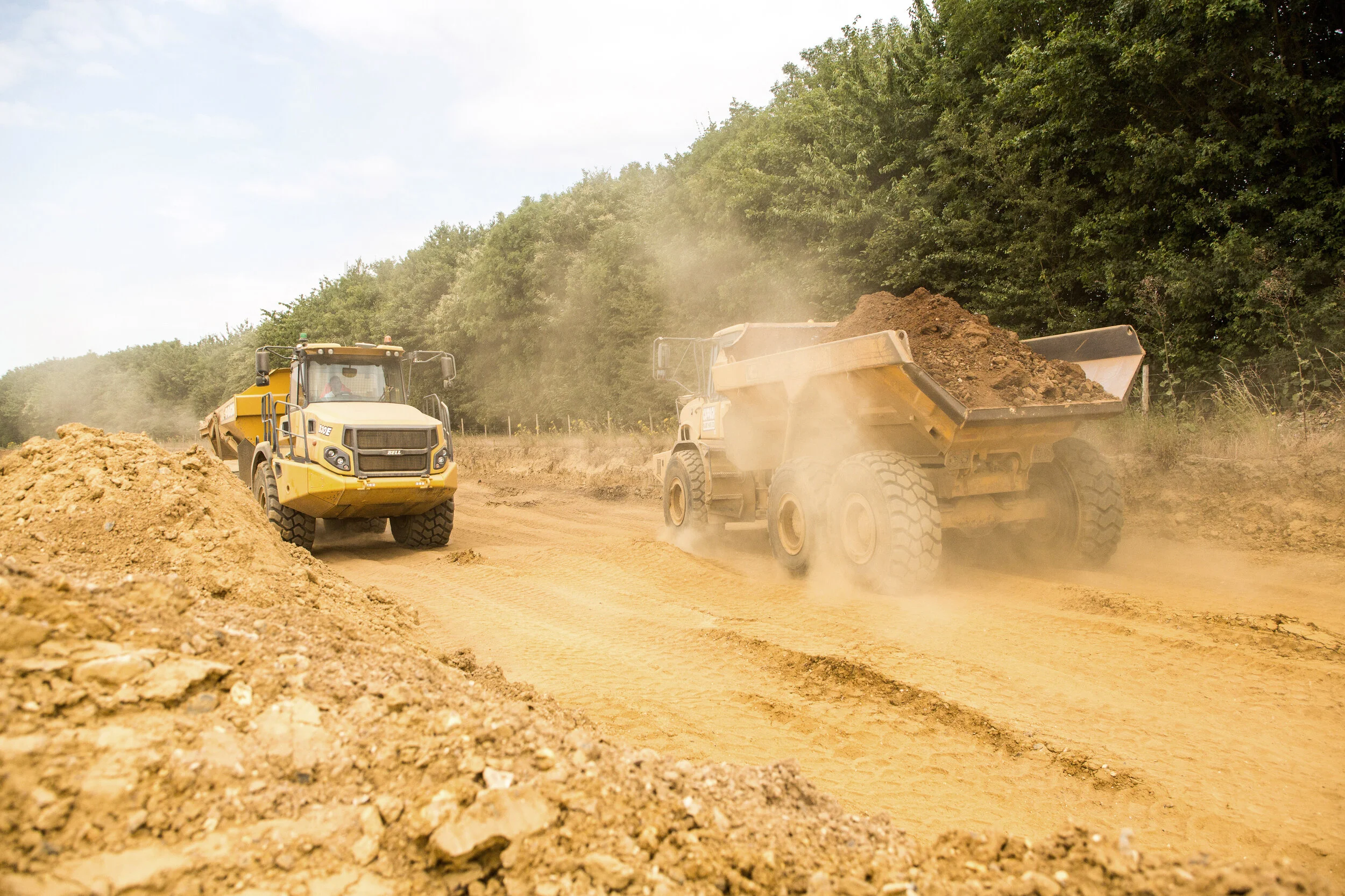 Construction site with two large dump trucks moving dirt and dust on a dirt road, surrounded by trees in the background.