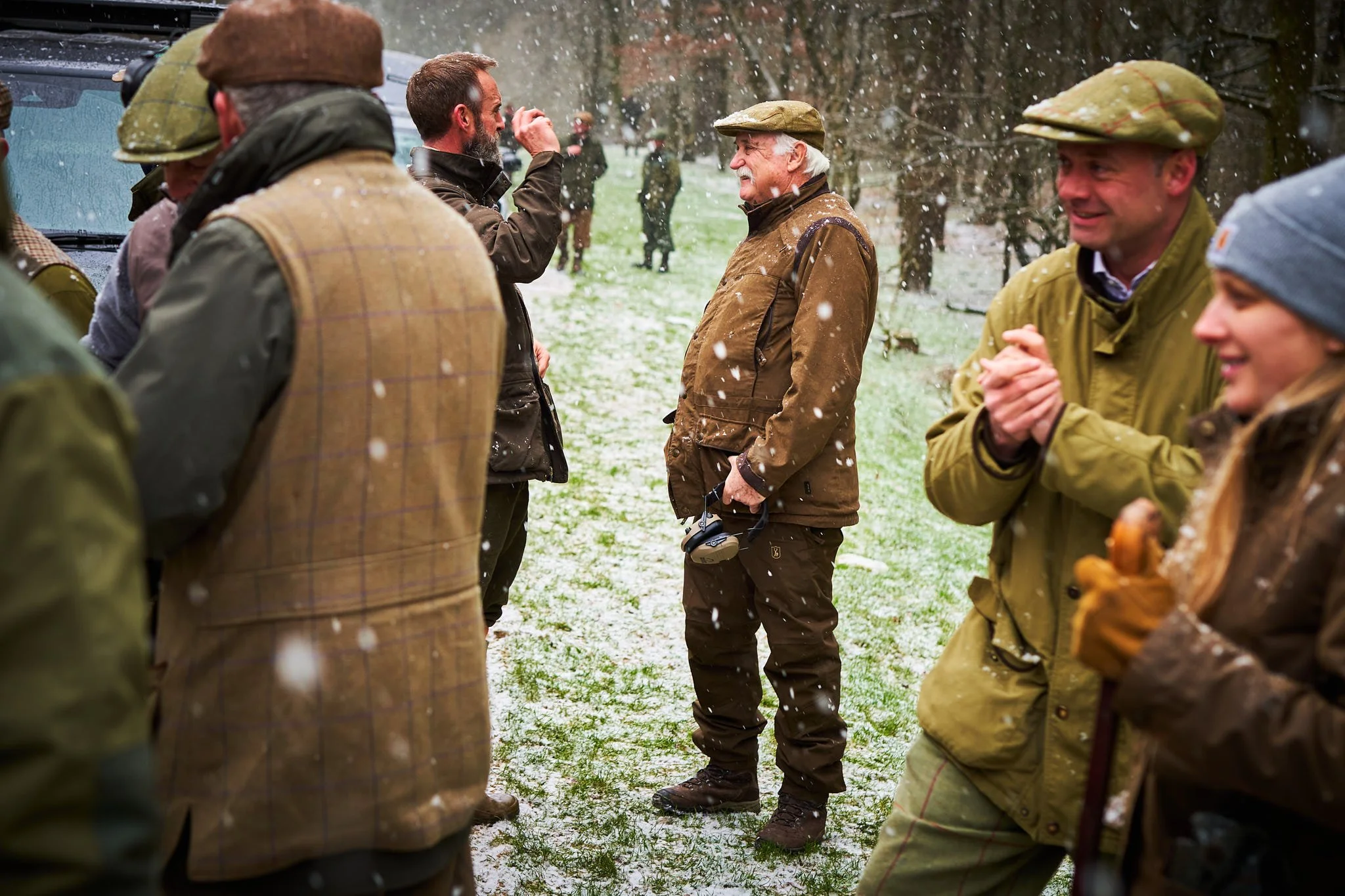 People dressed in outdoor and hunting attire, standing in a snowy field, smiling and engaging in conversation during a snow shower.