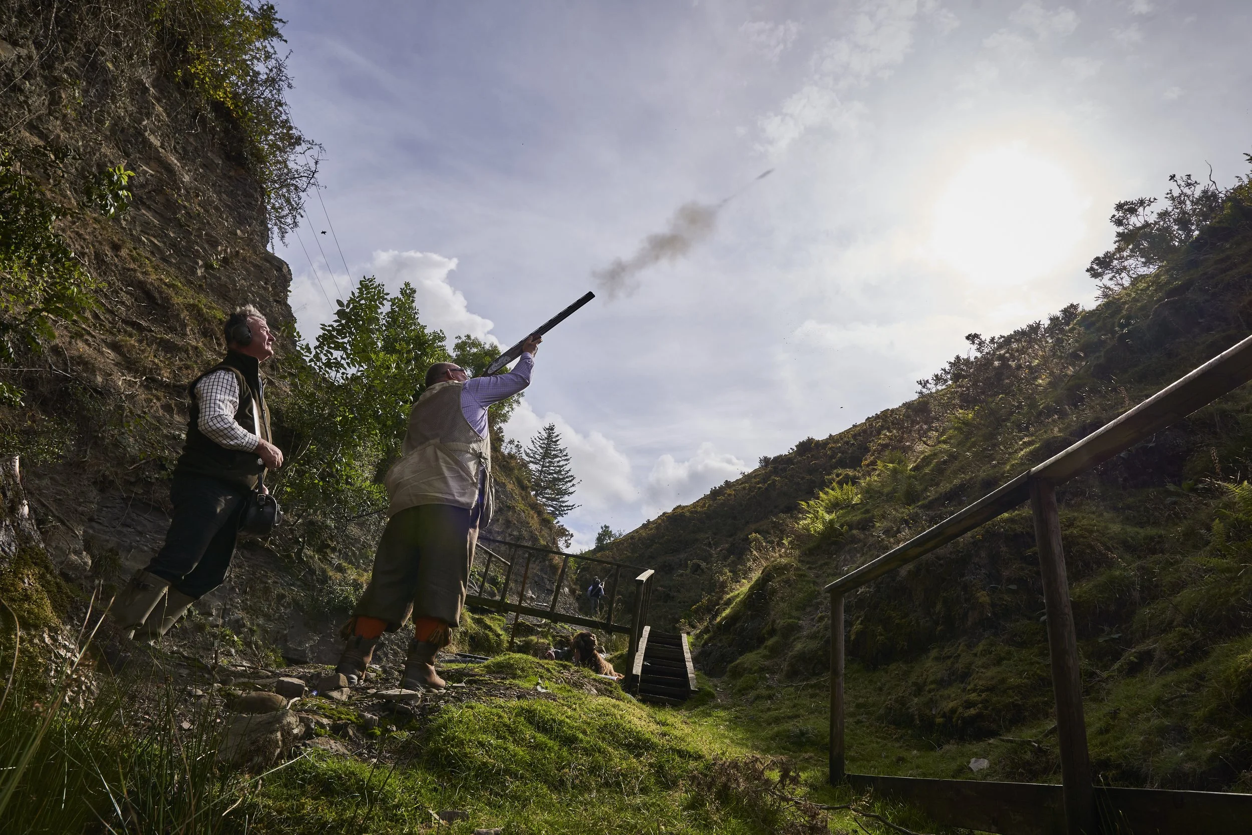 Two men in outdoor gear and boots releasing birdshot in a shotgun over a rocky hillside trail with green vegetation, a wooden railing, and a person crouching on the ground.