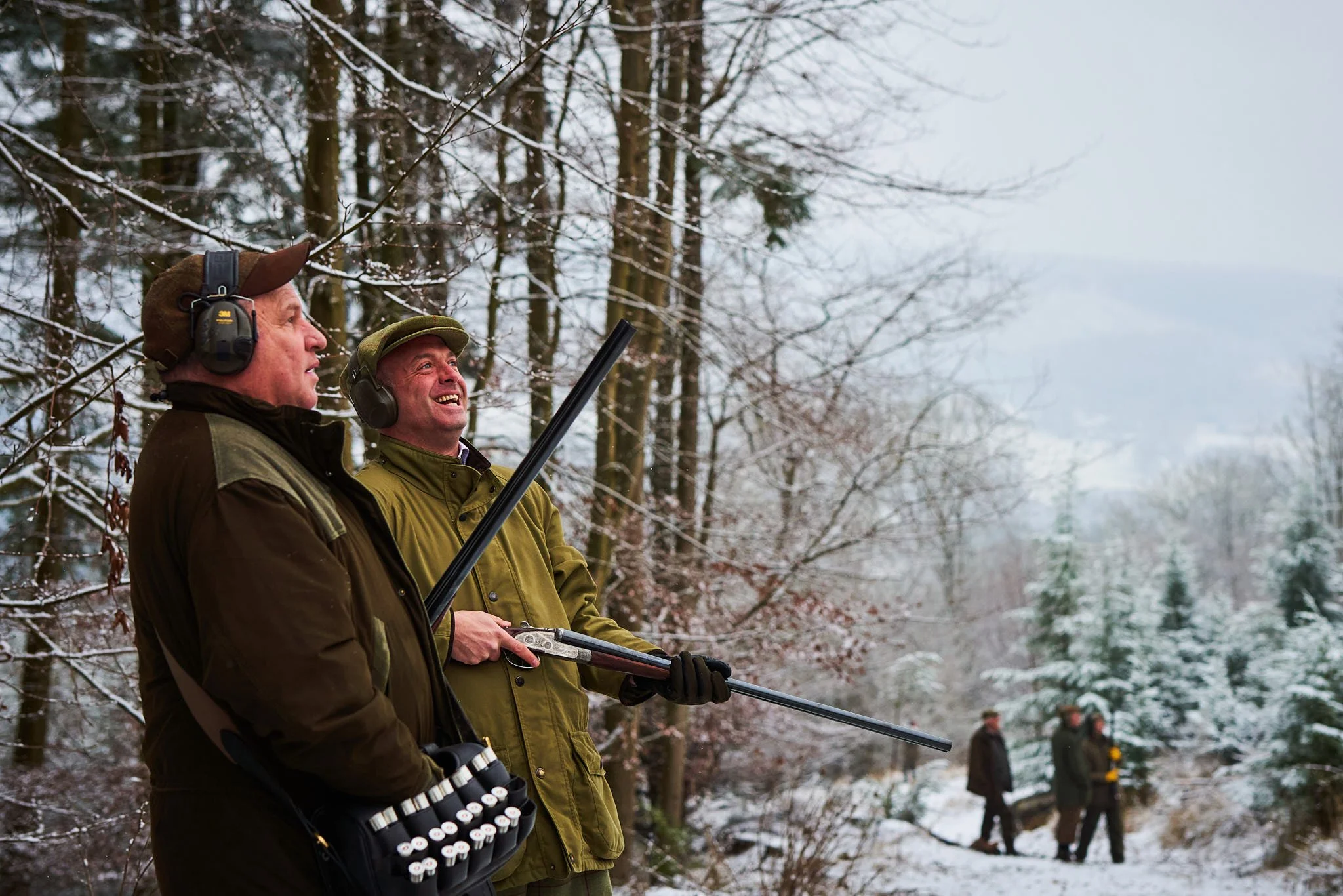 Two men outdoors in winter, wearing hunting gear and ear protection, holding a shotgun, standing in a snow-covered forest with other people in the background.