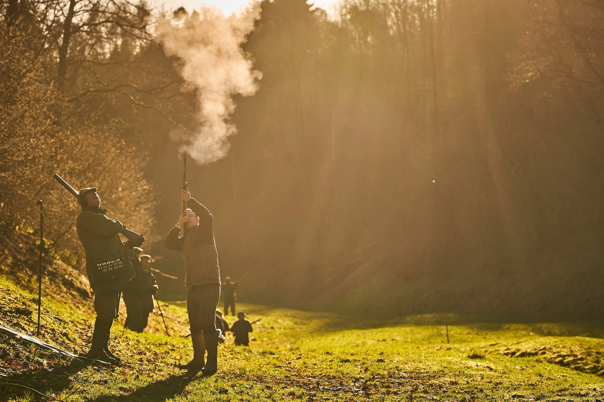 People outdoors in a wooded area during autumn, with sunlight filtering through the trees. One person is holding a smoke grenade, releasing smoke into the air, while others appear to be preparing or participating in an activity.