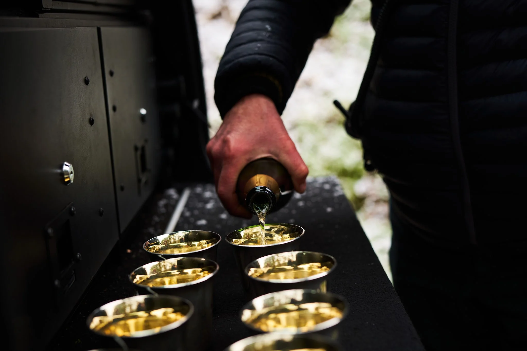 Person pouring champagne into several small glasses on a black table outdoors.