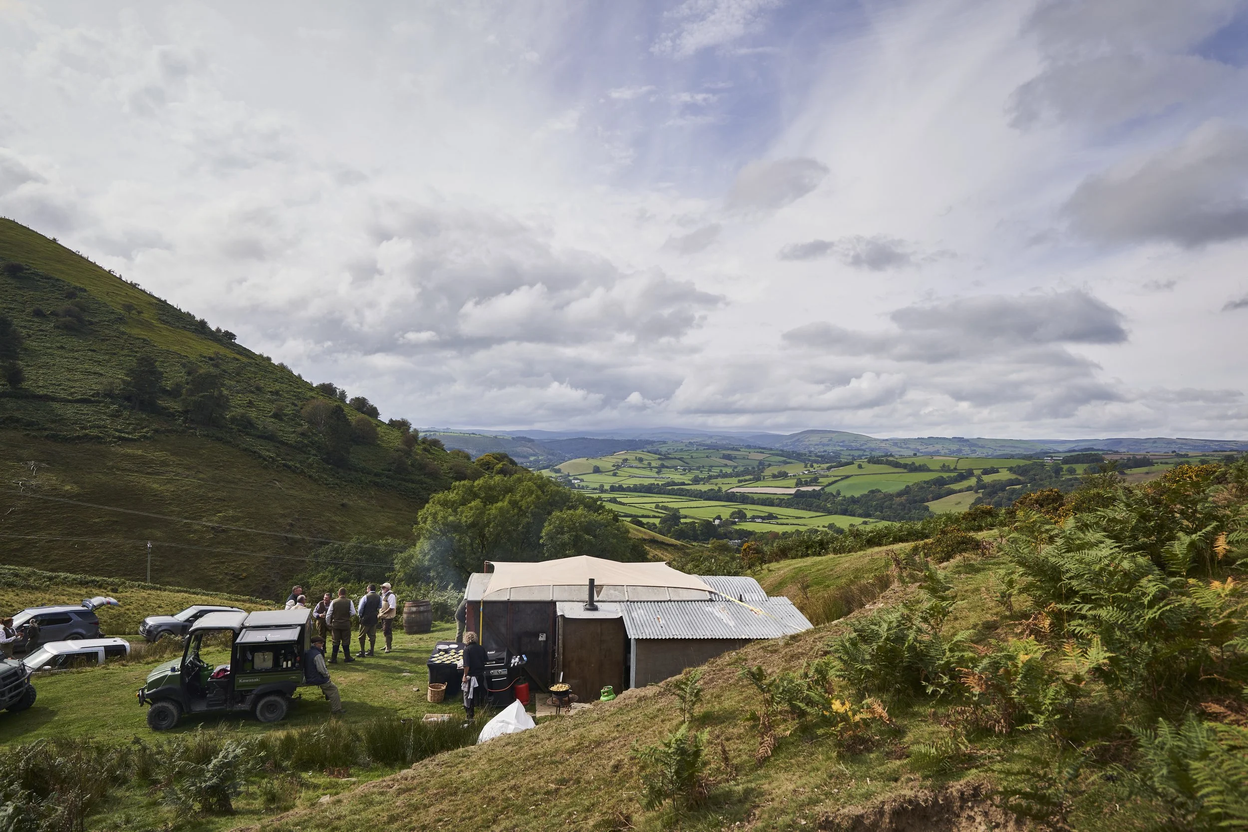 People gathered outdoors near a trailer on a grassy hillside with cars parked nearby and rolling green hills in the background under a cloudy sky.
